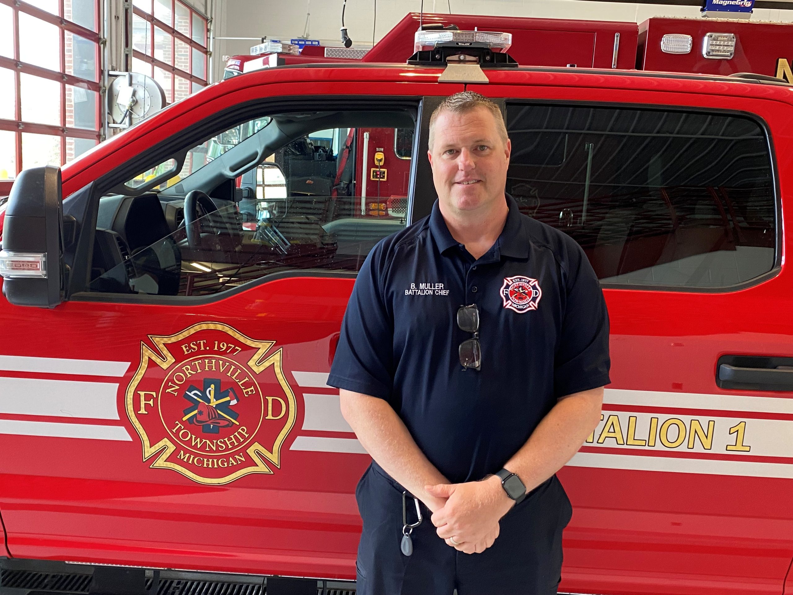 Northville Township Fire BC Brent Muller - Social House News Fire Battalion Chief B. Muller stands in front of a red fire engine inside a fire station in uniform.