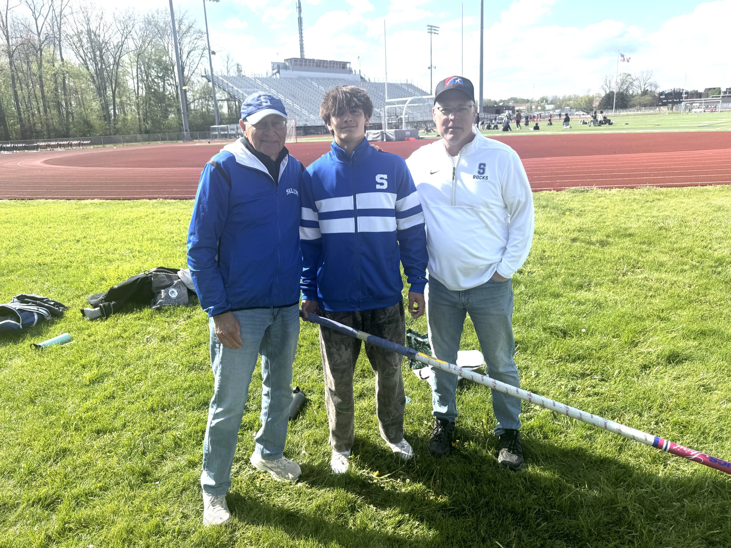 Three men stand together on a grassy track field, wearing blue and white jackets, posing for a photo with a sports stadium in the background.