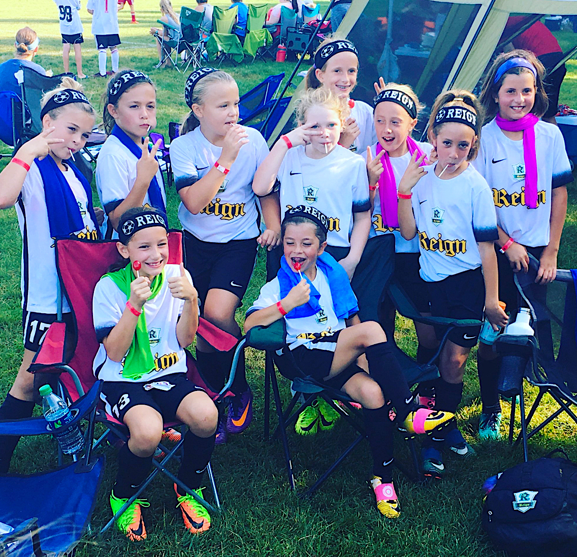 Group of young girls in white and black soccer uniforms posing for a team photo at a park camp, smiling and making peace signs; tents and chairs in the background indicate a tournament or event.