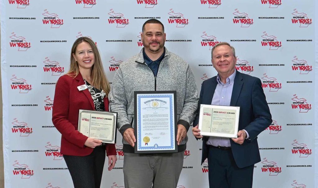 Three individuals posing with awards; woman in a red blazer on the left, center man with a large framed certificate, and man in a suit on the right, in front of a Michigan Works! backdrop.