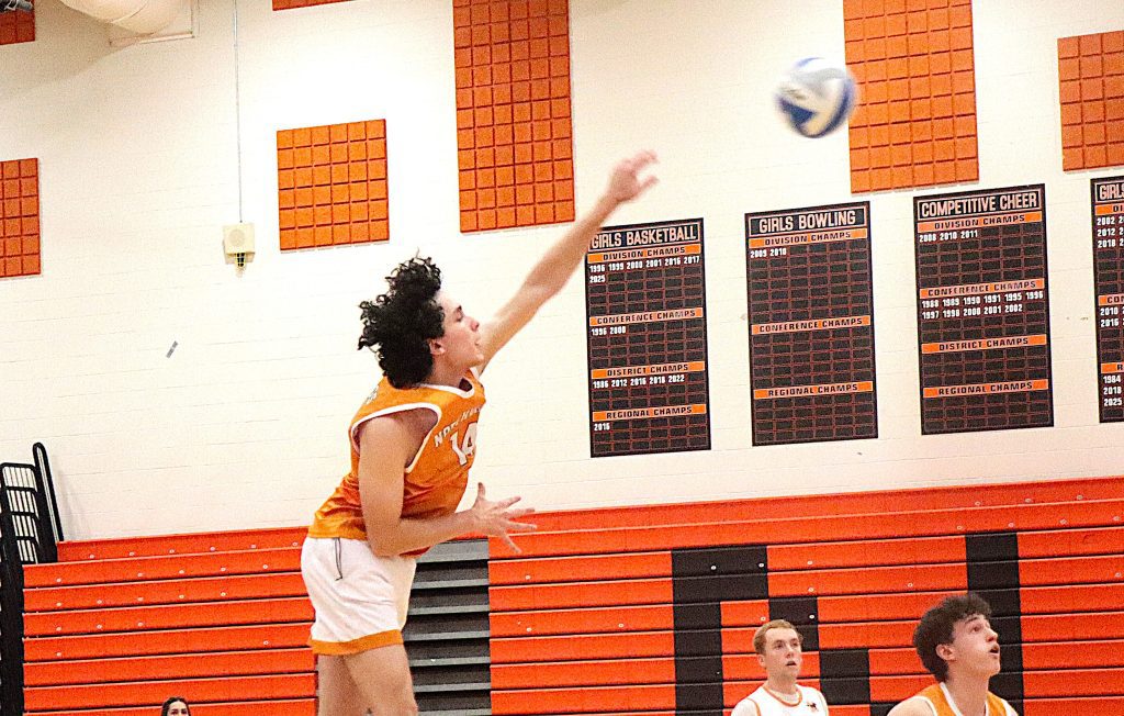 Girl in orange jersey jumps to hit a ball in a school gym with orange bleachers and wall banners behind her.