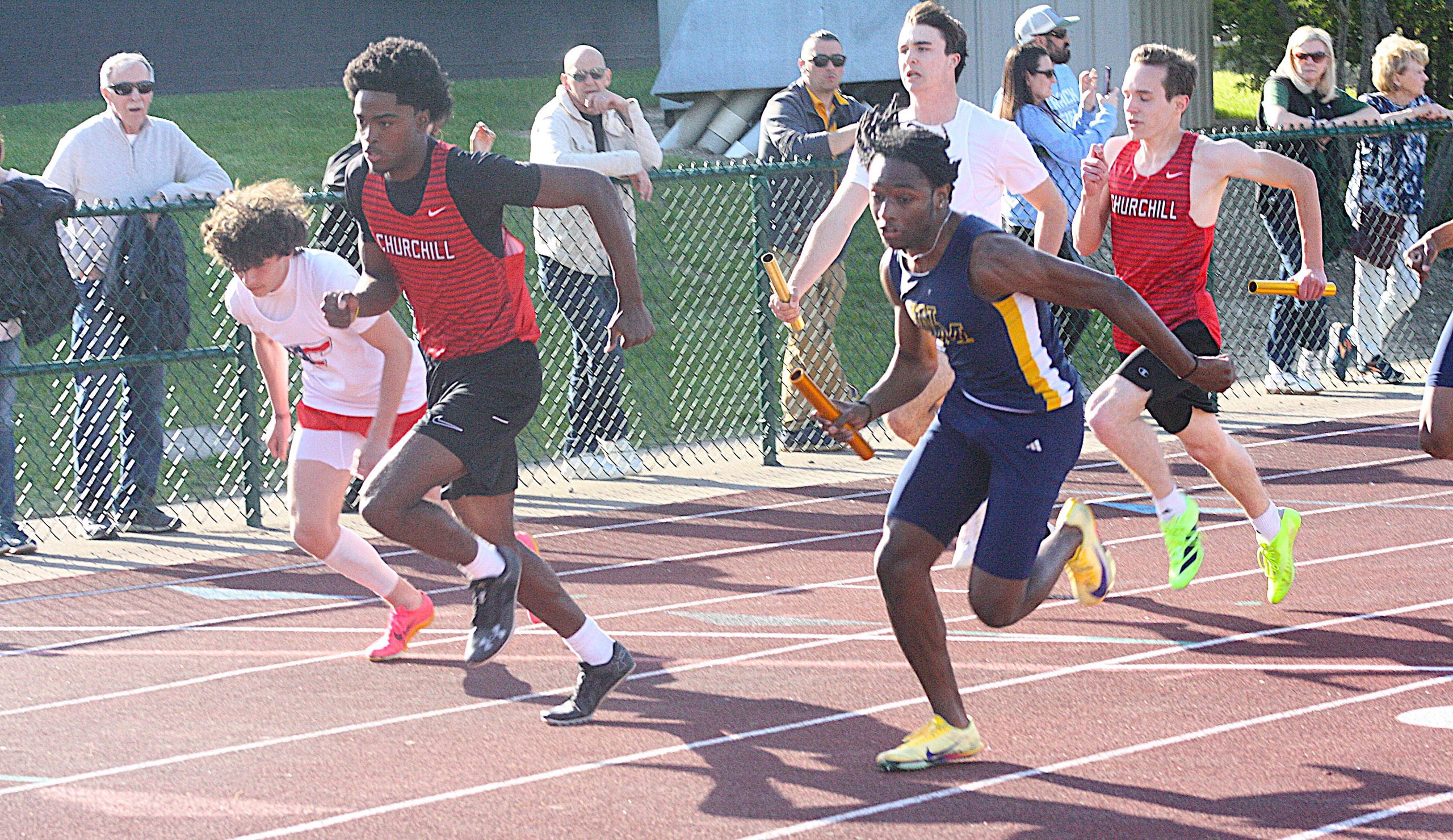 TFreg1 - Social House News Several male sprinters race on a track, wearing Churchill red and blue uniforms, with spectators watching behind a chain-link fence.
