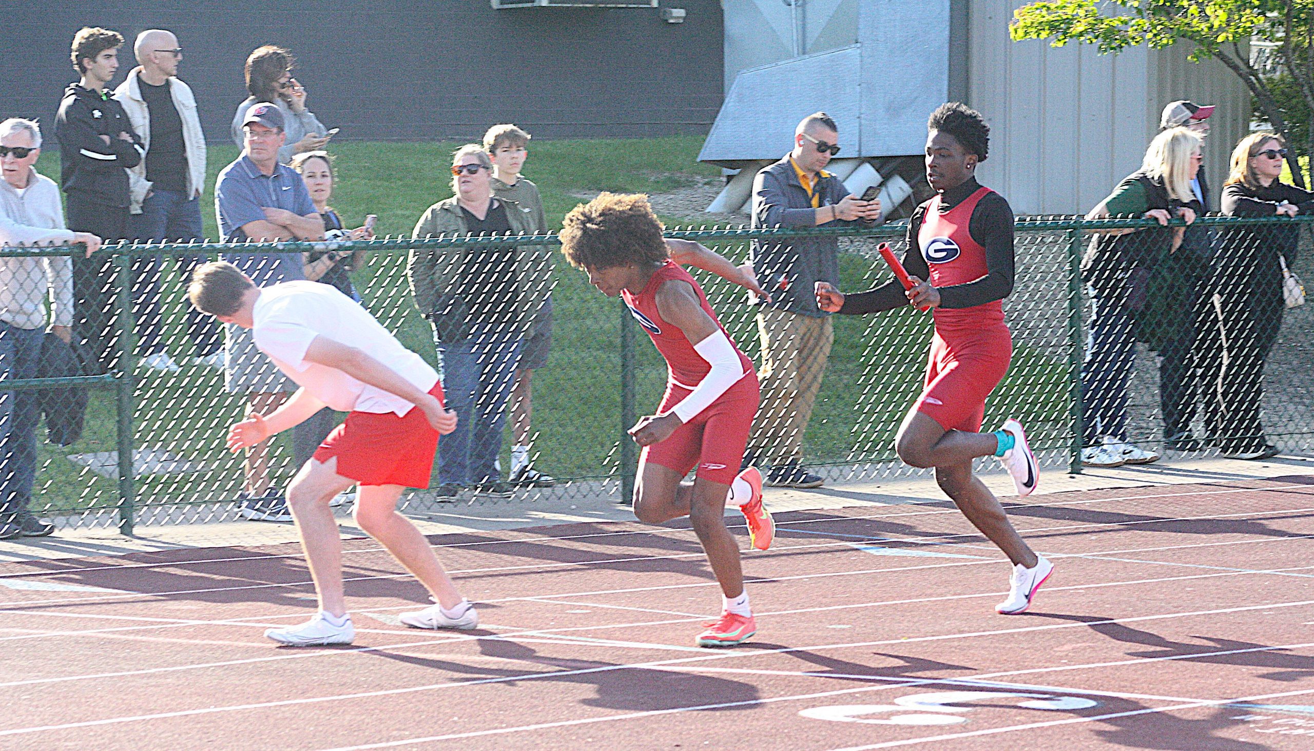 TFreg2 - Social House News Runners in red uniforms sprint on a track while spectators watch from behind a chain-link fence.