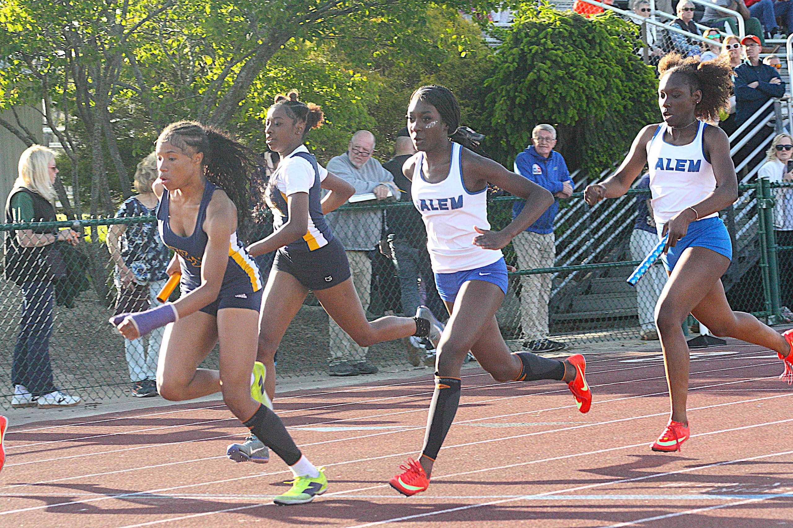 TFreg3 - Social House News Group of female sprinters racing on an outdoor track, wearing blue and white uniforms with 'ALEM' on their jerseys, behind a chain-link fence with spectators beyond.