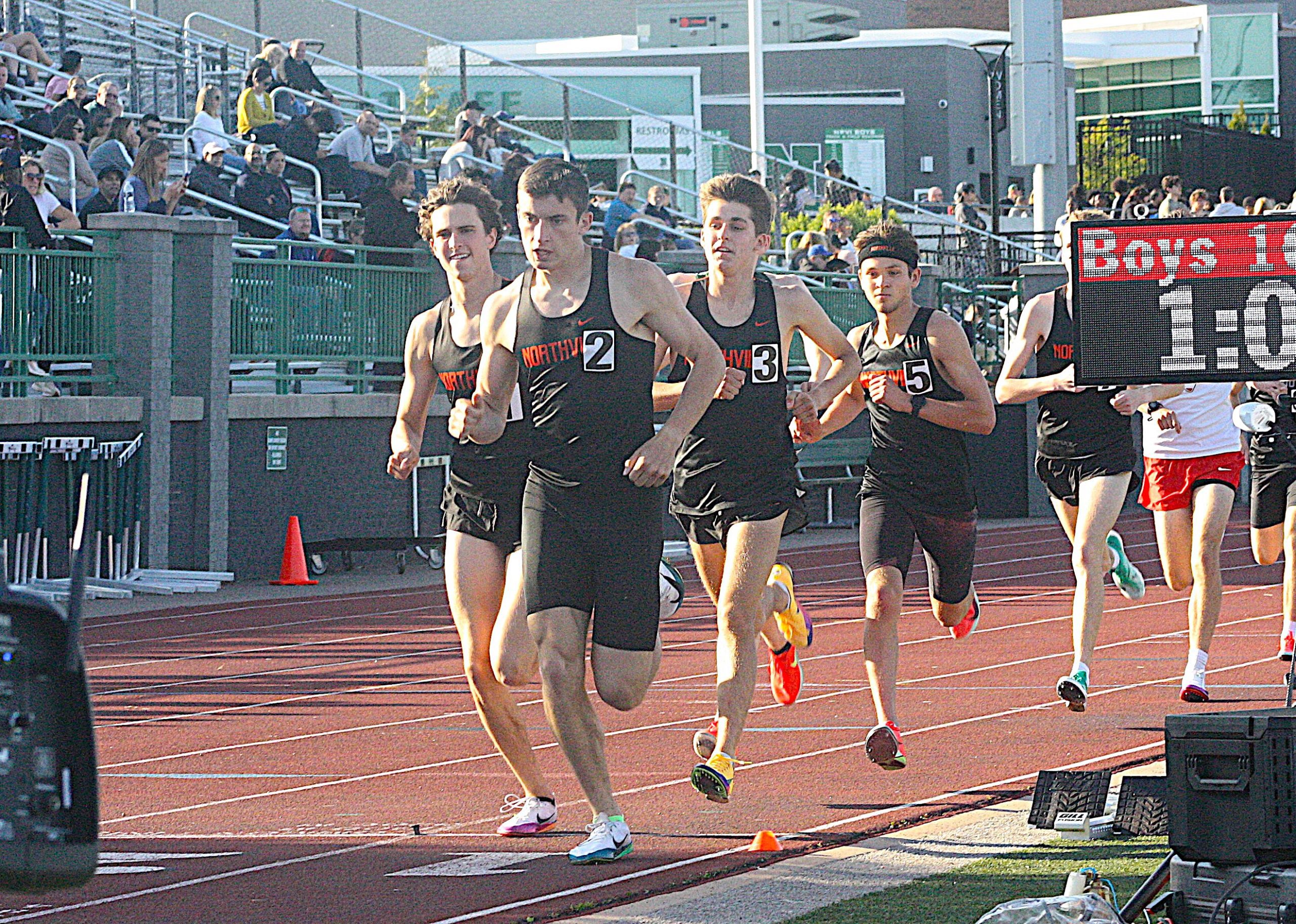 TFreg4 - Social House News Four male sprinters in black uniforms race on a red track as spectators watch from the stands.
