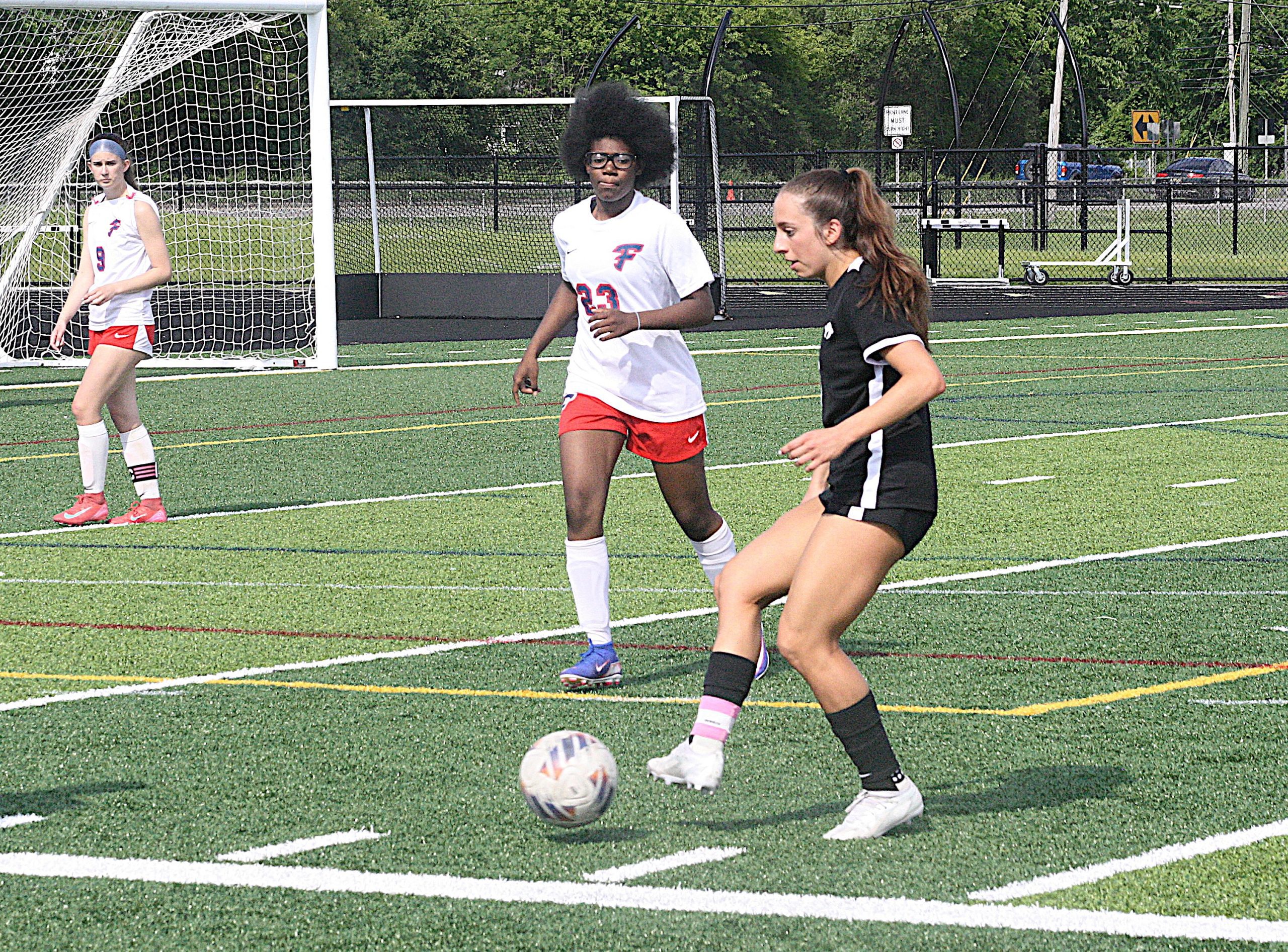 alik10 - Social House News Soccer players in white and black uniforms chase a ball on a turf field near a goal net, with one player in a black kit in the foreground.