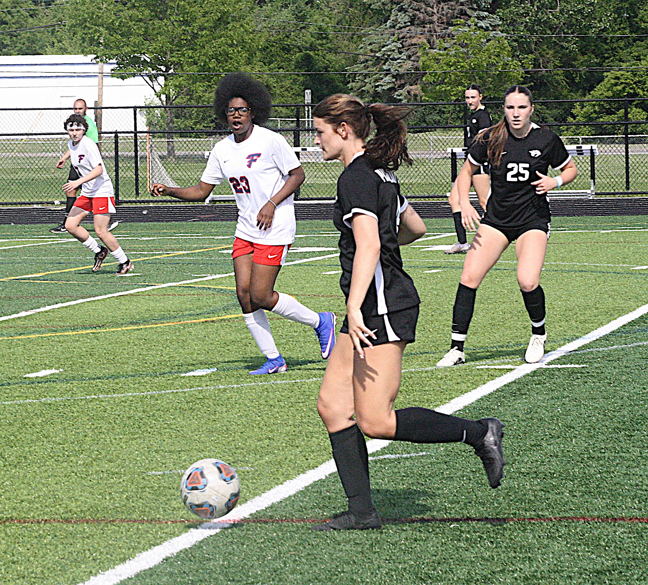 Young soccer players chase a ball on an outdoor turf field, wearing white and black uniforms with a chain-link fence and trees in the background.