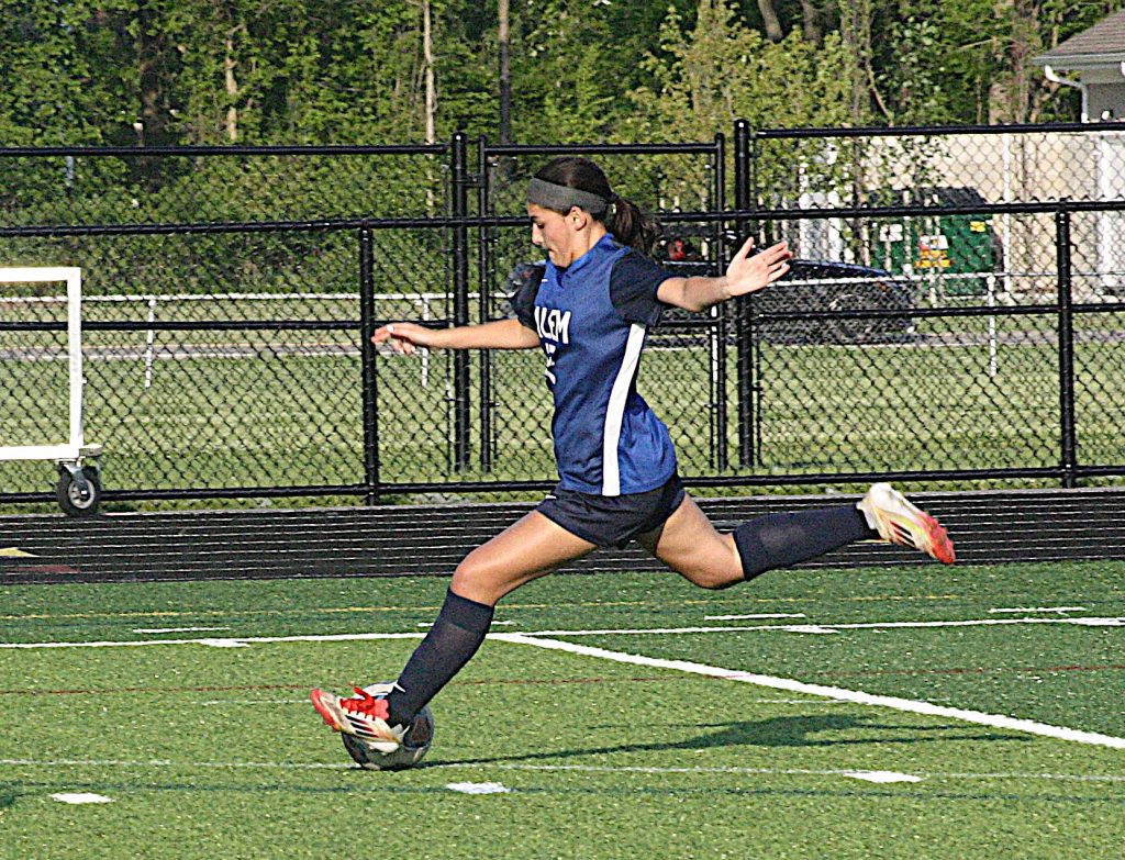 Female soccer player in blue jersey and black shorts kicking a ball on a turf field, mid-stride with arms out for balance.
