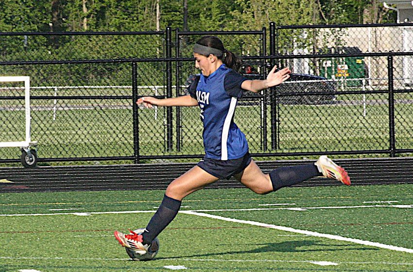 Female soccer player in blue jersey and black shorts kicking a ball on a turf field, mid-stride with arms out for balance.