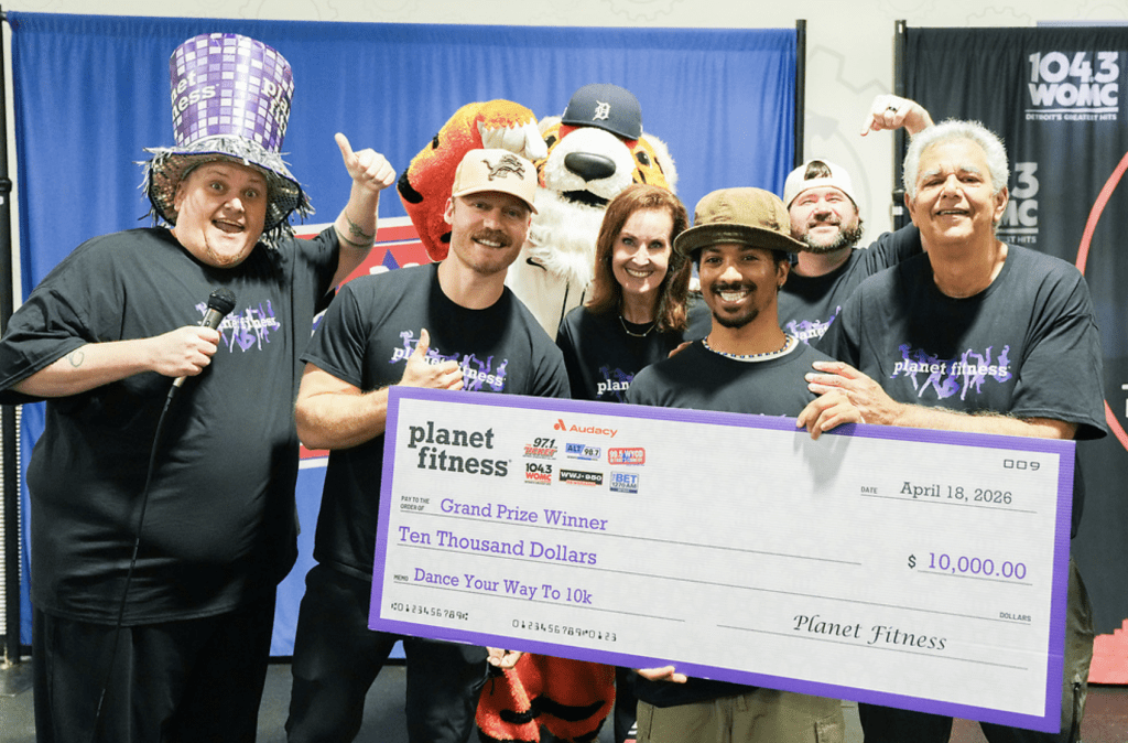 Group of people in black Planet Fitness shirts pose with an oversized check for $10,000 at a charity event, smiling at the camera.