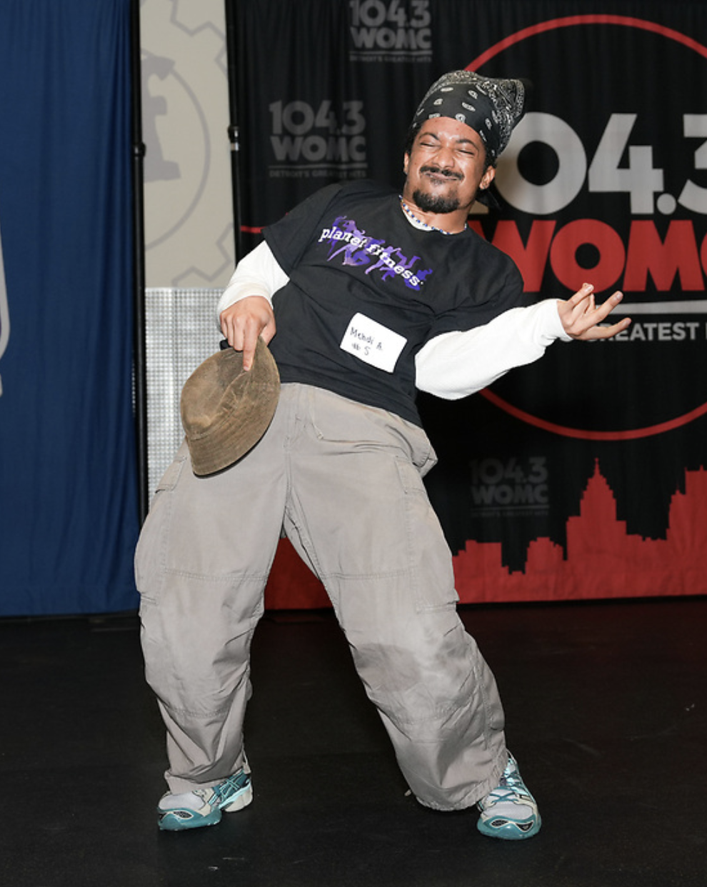 Male breakdancer wearing a bandana, black shirt, gray cargo pants, turquoise sneakers, holding a hat mid-move on stage.