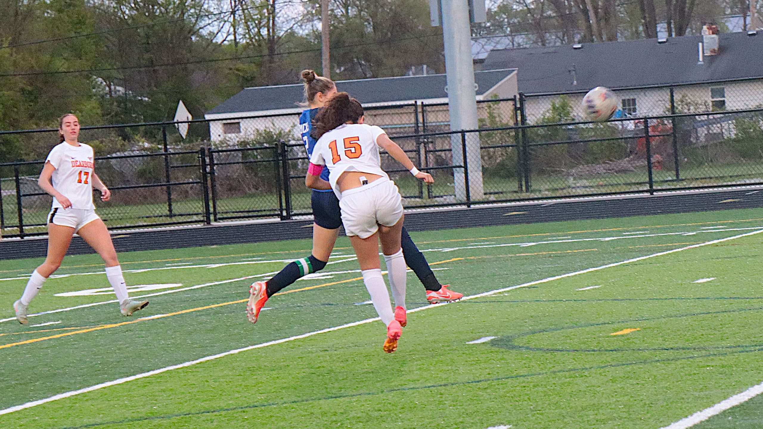 Soccer players in white uniforms challenge for the ball on a green artificial turf field, with a ball midair to the right and a fence in the background.