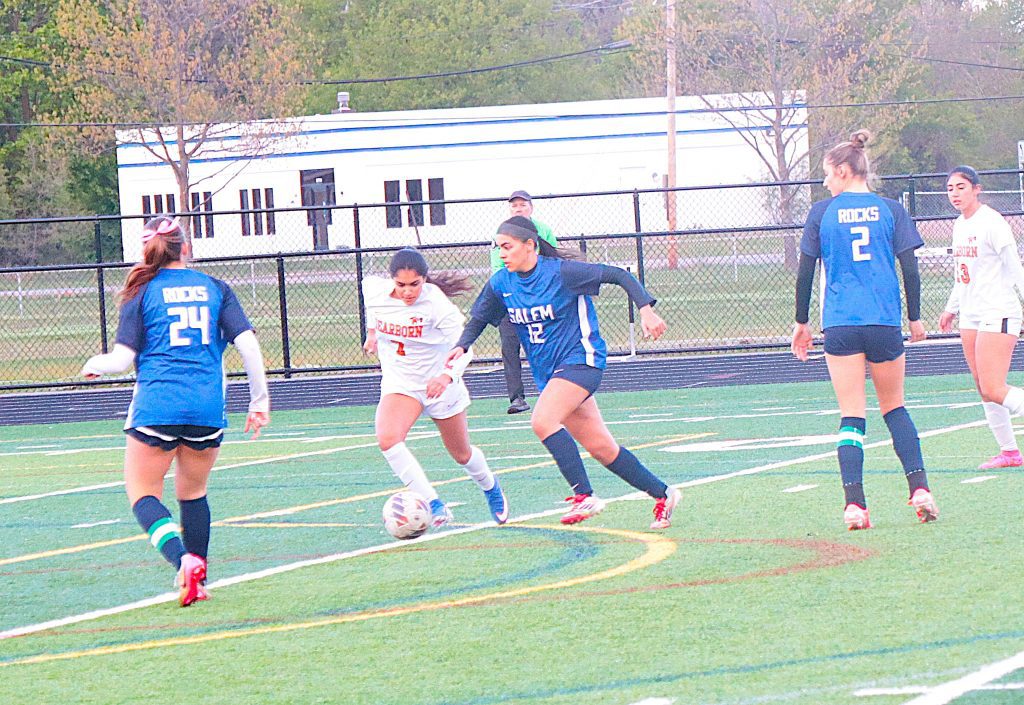 Girls' soccer match on a turf field; players in blue and white jerseys chase a ball near the center, with a chain-link fence and trees in the background.
