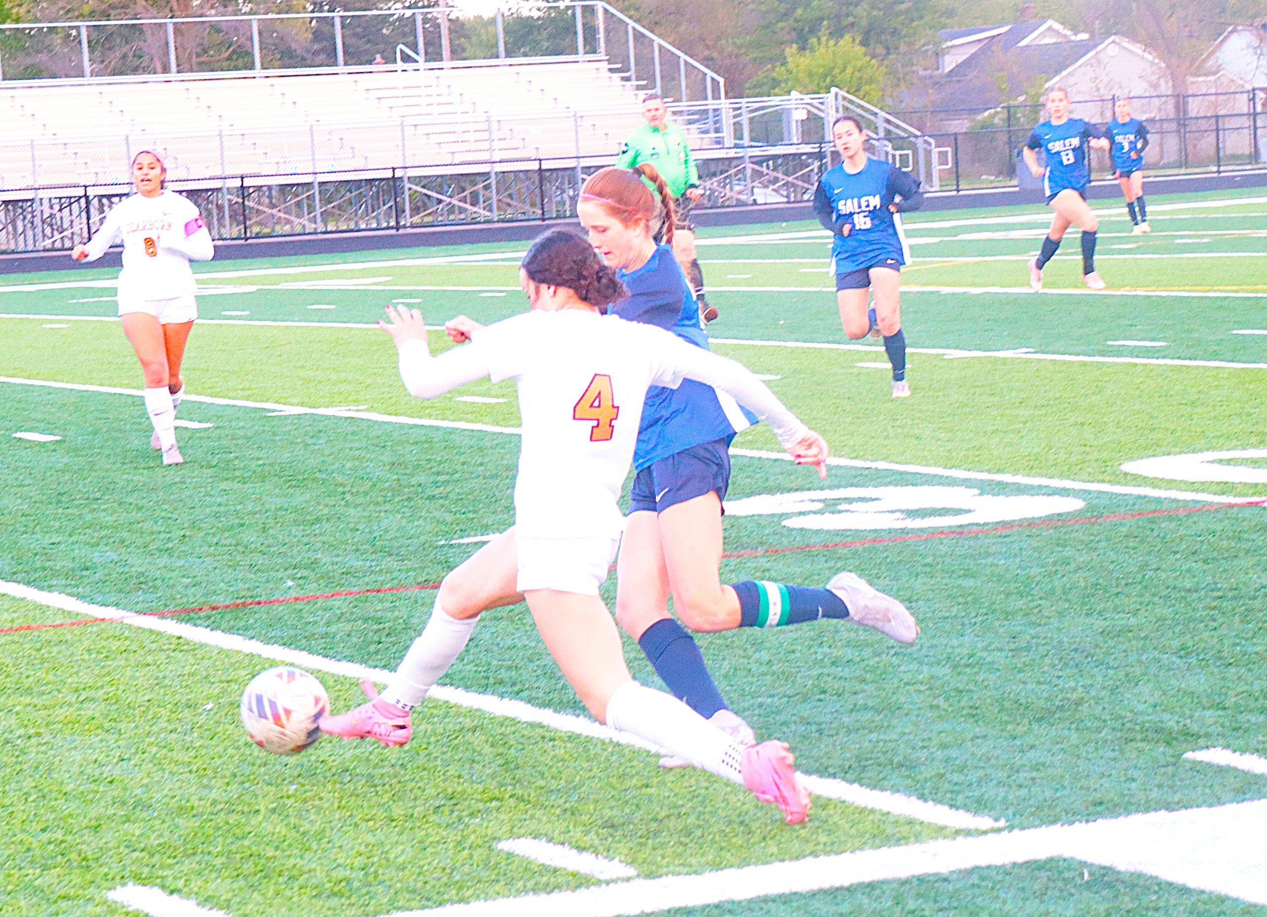 Girls’ soccer match in progress: foreground players in white jersey (number 4) and blue jerseys chase the ball on a green turf field with bleachers in the background.