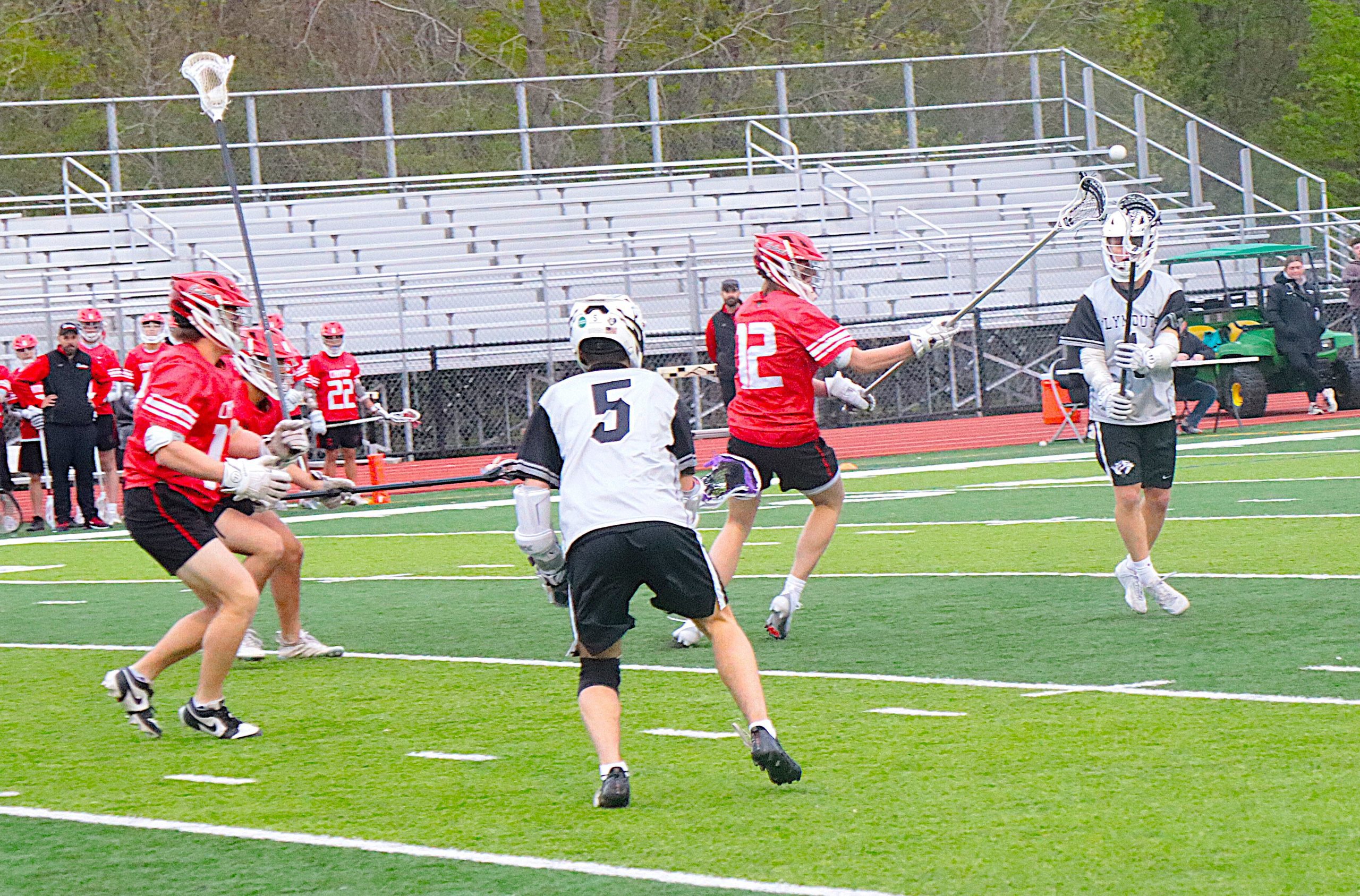 Lacrosse players in red and white uniforms race for the ball on a green turf field during a game, with bleachers in the background.