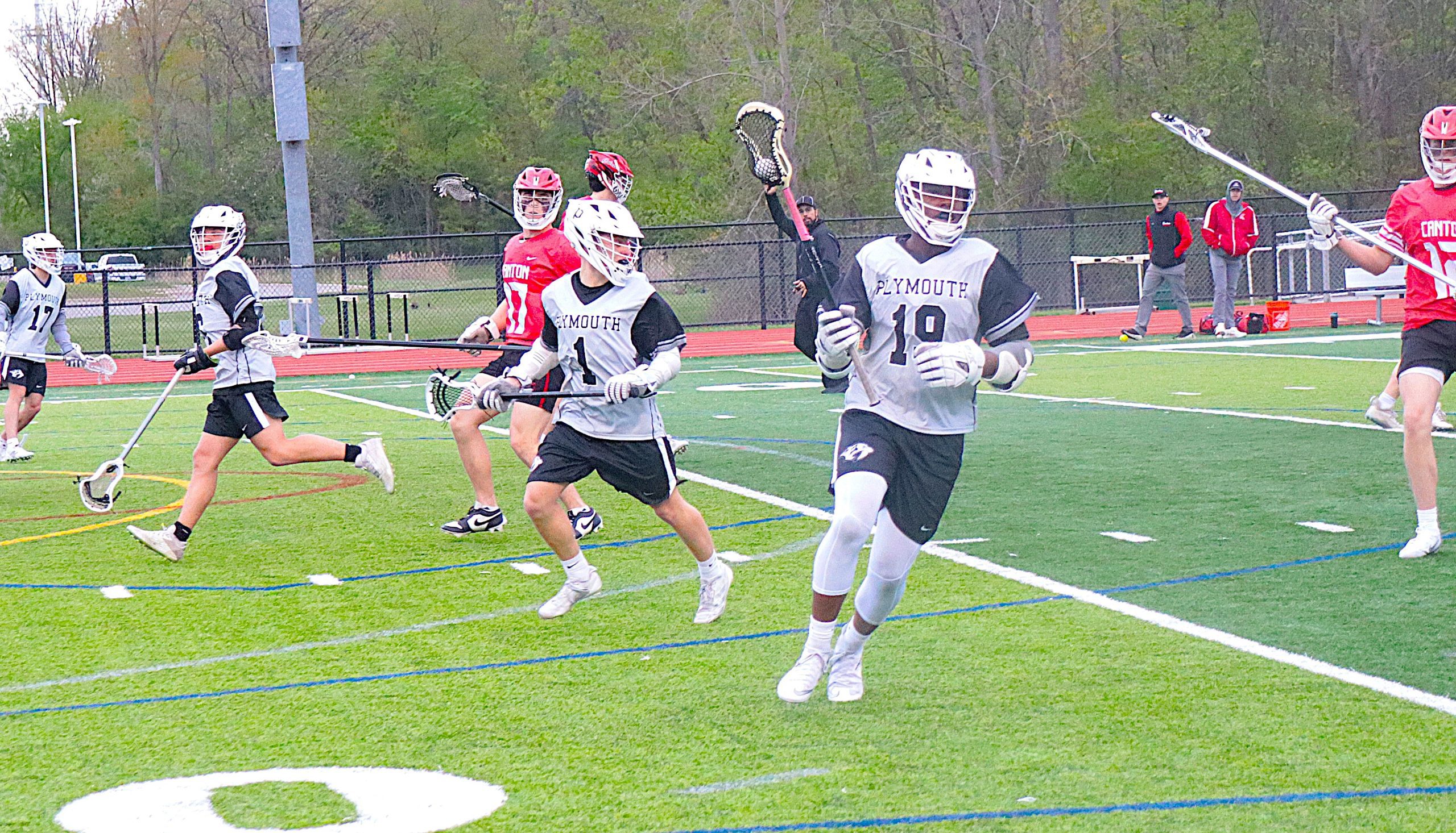 Lacrosse players sprint across a turf field during a game, white and gray jerseys contrasting with red opposing uniforms.