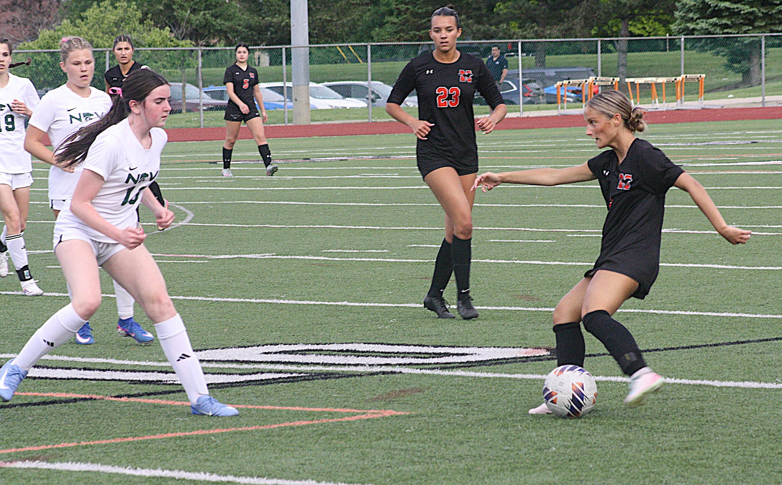 nvnovisoc1 - Social House News Girls' soccer match on artificial turf with players in white and black uniforms; ball near a player in black jersey #23 and a teammate in white ahead.