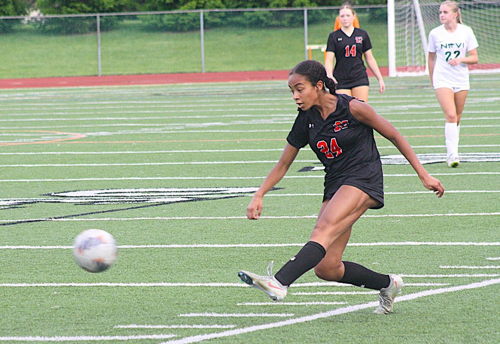 Female soccer player in a black uniform (#24) kicking a ball on a green field during a match, with opposing players in the background.