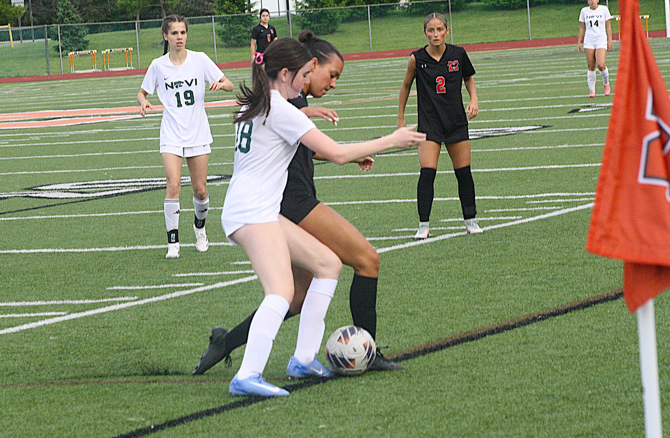nvnovisoc3 - Social House News Girls' soccer players in white and black jerseys scramble for the ball near the sideline by the corner flag on a turf field from a side angle.