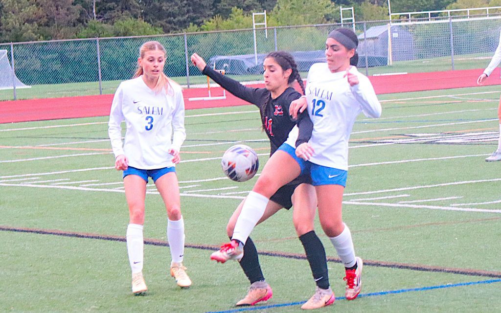 Three female soccer players vie for the ball on a green turf field; two wear white Salem jerseys (#3 and #12) flanking a Black jersey player (#11).