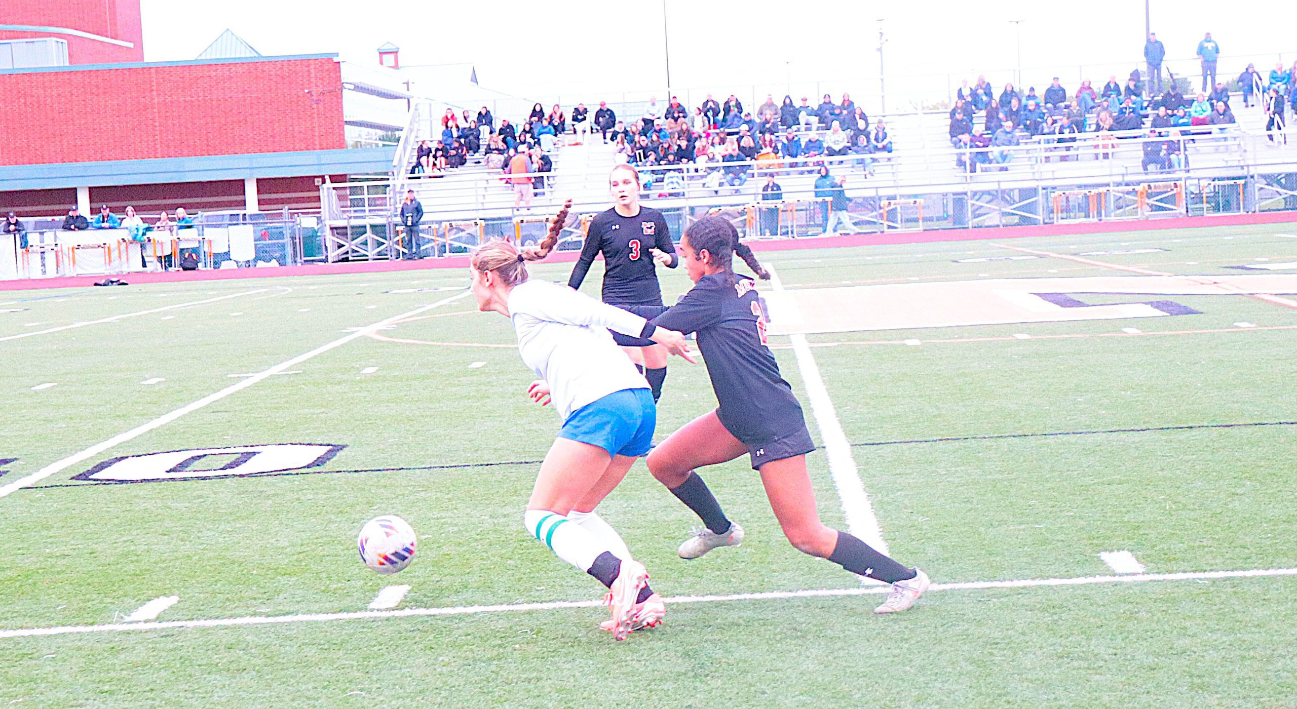 Soccer players in white and black uniforms compete for the ball on a turf field while spectators watch from the stands behind them.