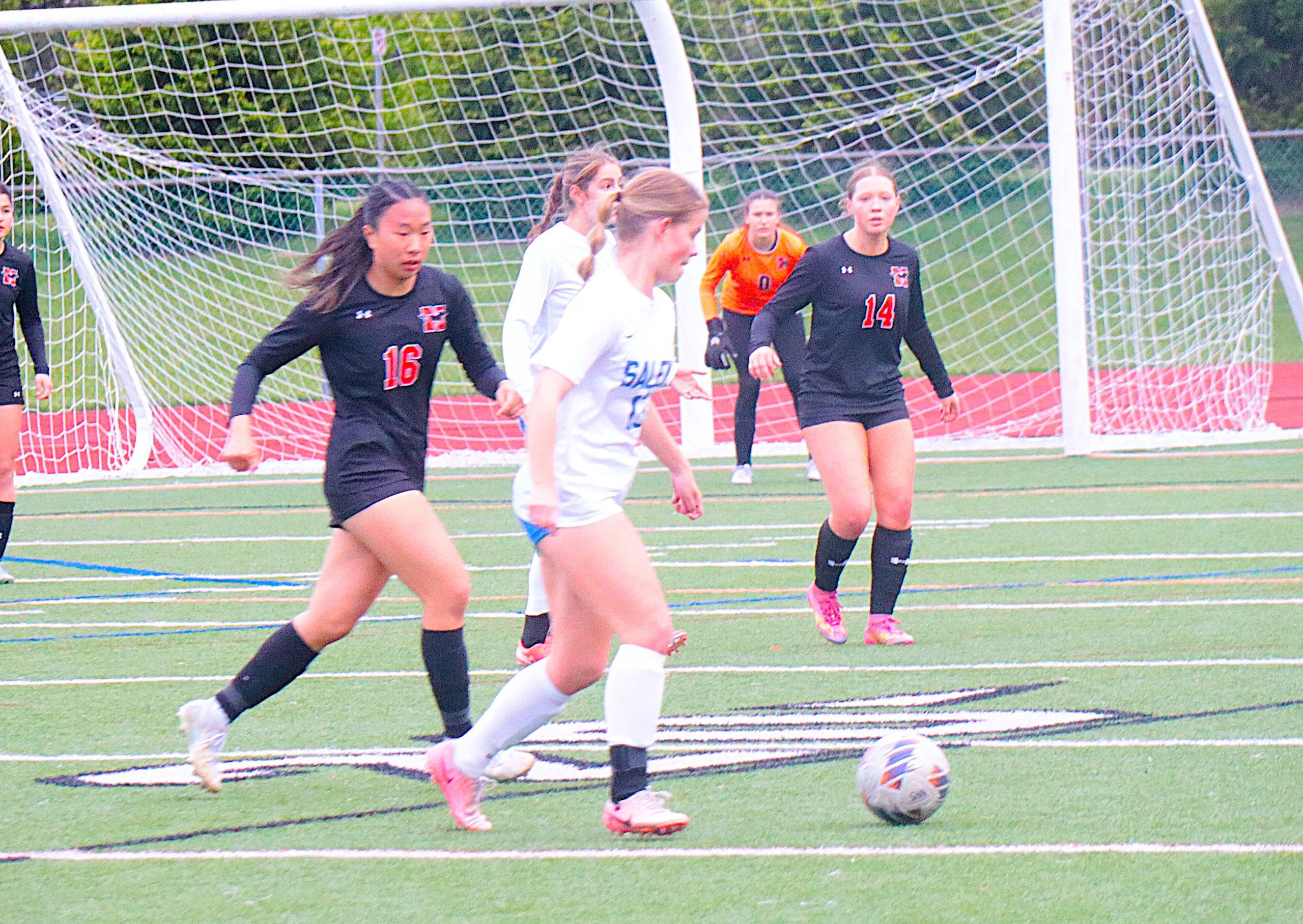 Soccer players chase the ball near a goal on a green turf field, with a goalkeeper in orange in the background.