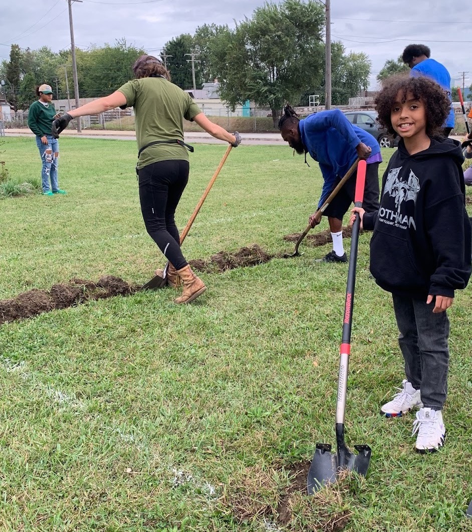 Group of young volunteers digging holes in a grassy field for a community project; a girl in the foreground holds a shovel and smiles at the camera.