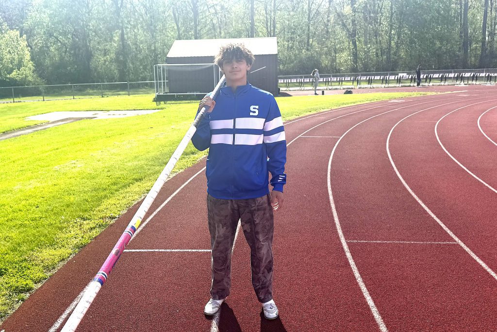 Young person on a red running track holding a long pole vault pole, wearing a blue jacket with white stripes on the chest.