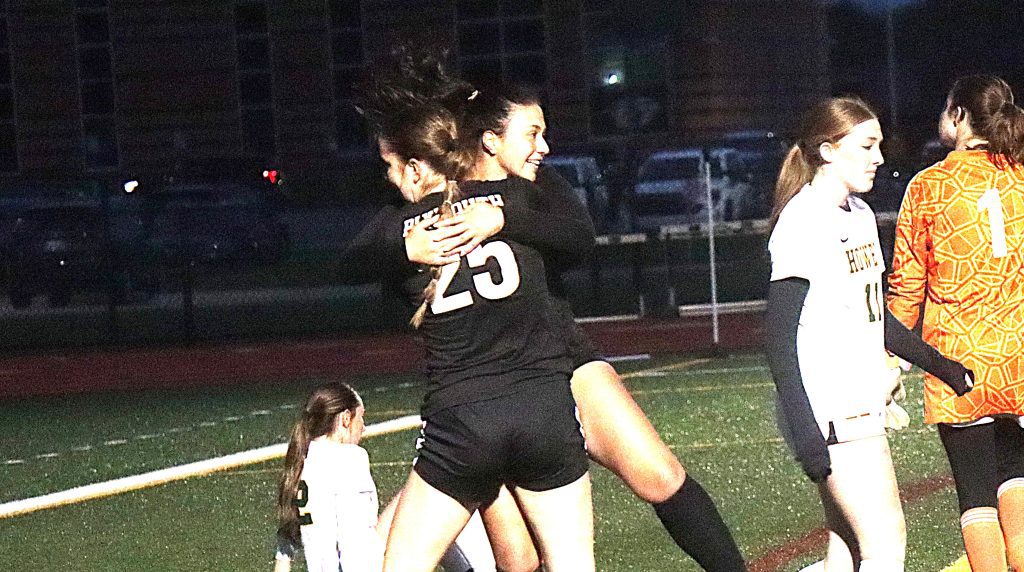 Two players in black uniforms hug in celebration on a night-time sports field, with teammates nearby in white and orange jerseys.