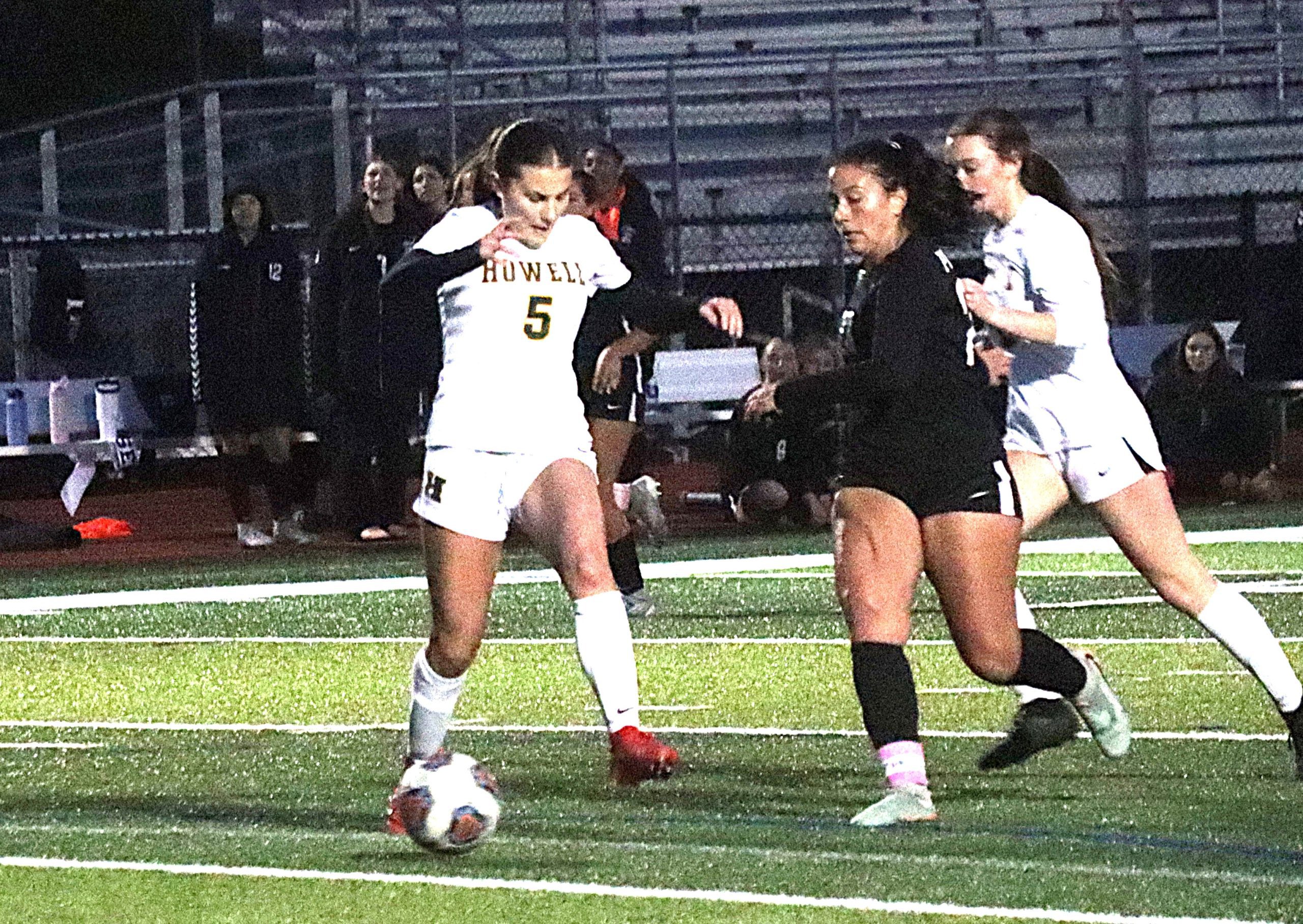Soccer players chase a ball on a lit green field during a night game; white and black uniforms in motion.