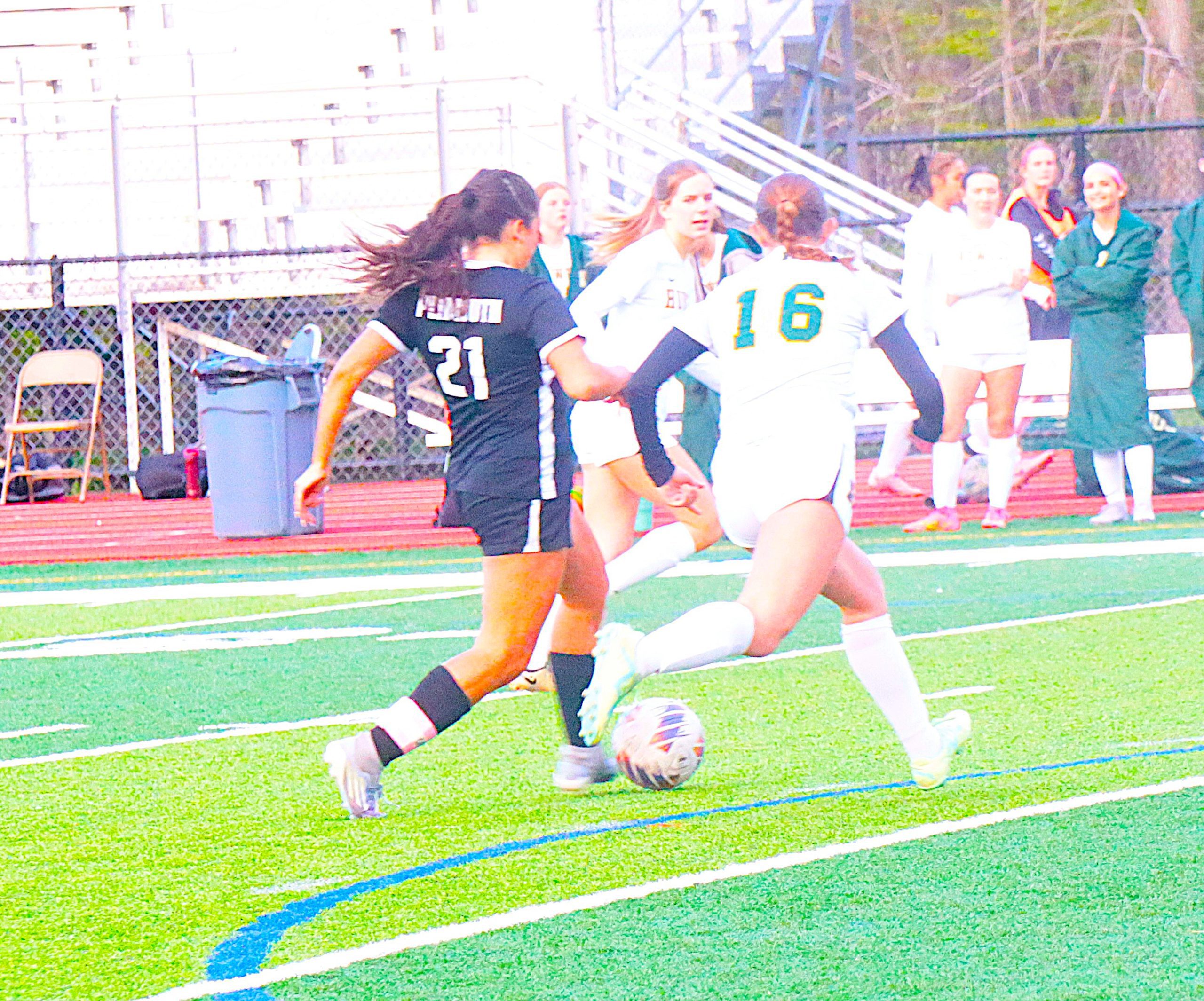 Girls' soccer game on green turf; player in white jersey (number 16) dribbles near the ball while others pursue.