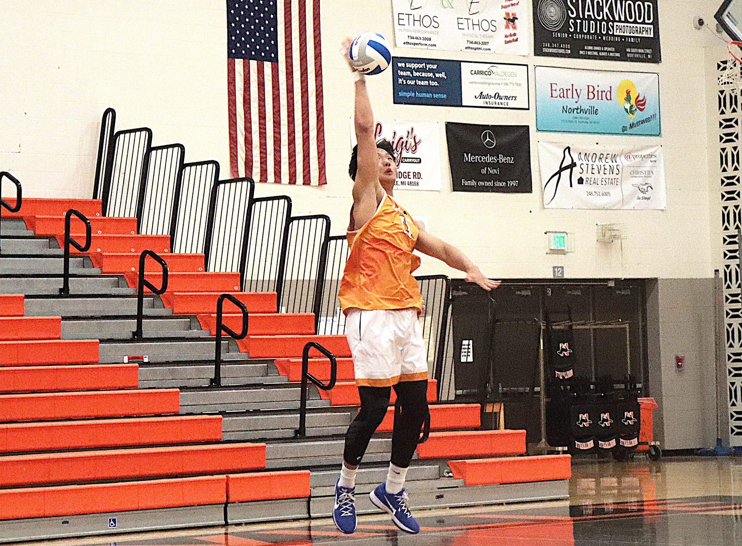 Volleyball player in orange jersey leaps to strike a ball in a gym, with orange bleachers, an American flag, and wall banners in the background