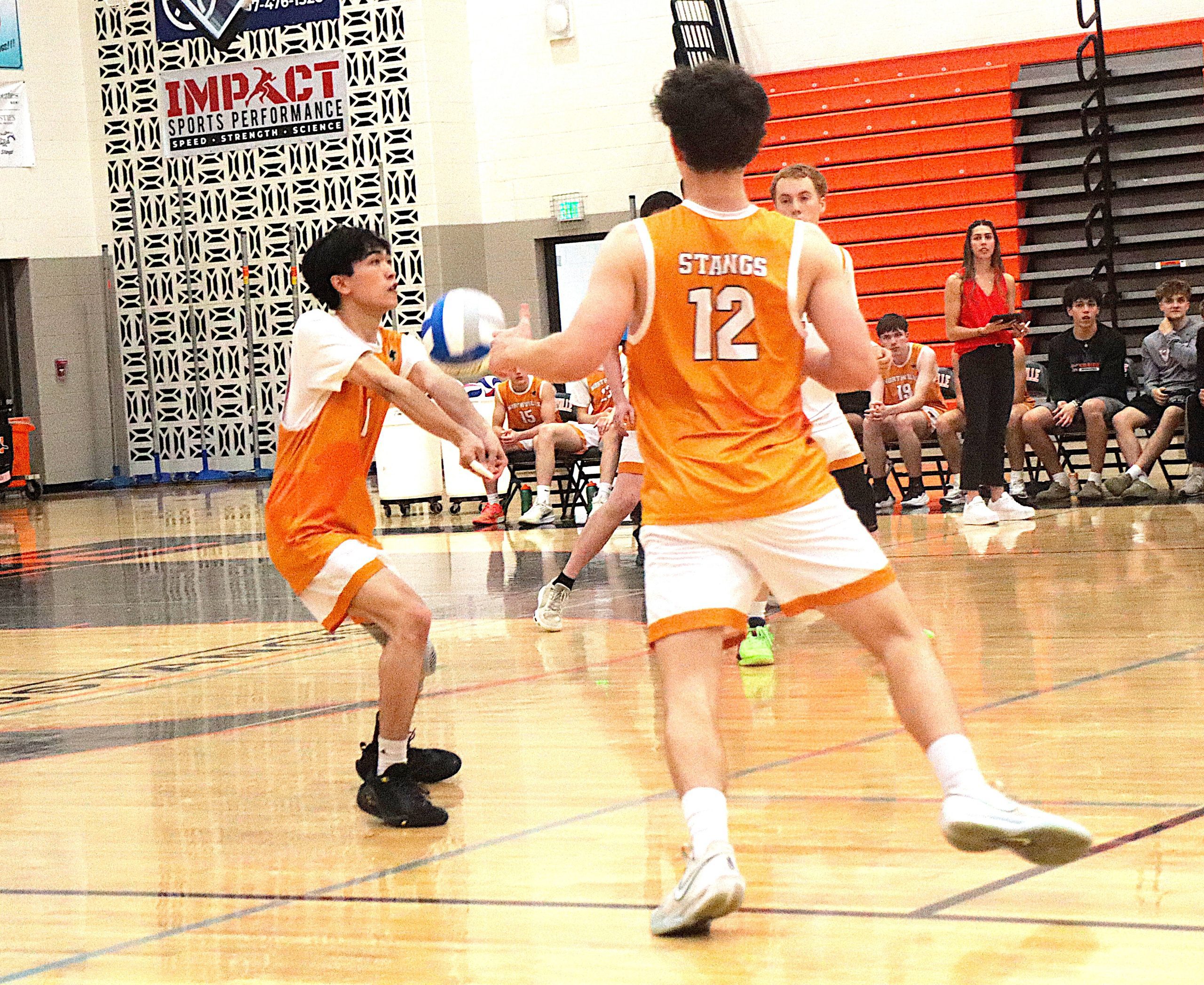 Two players in orange uniforms scramble for a basketball on a gym floor, one with his back to the camera wearing jersey #12 as teammates and spectators look on in the background and orange bleachers rise on the right side.
