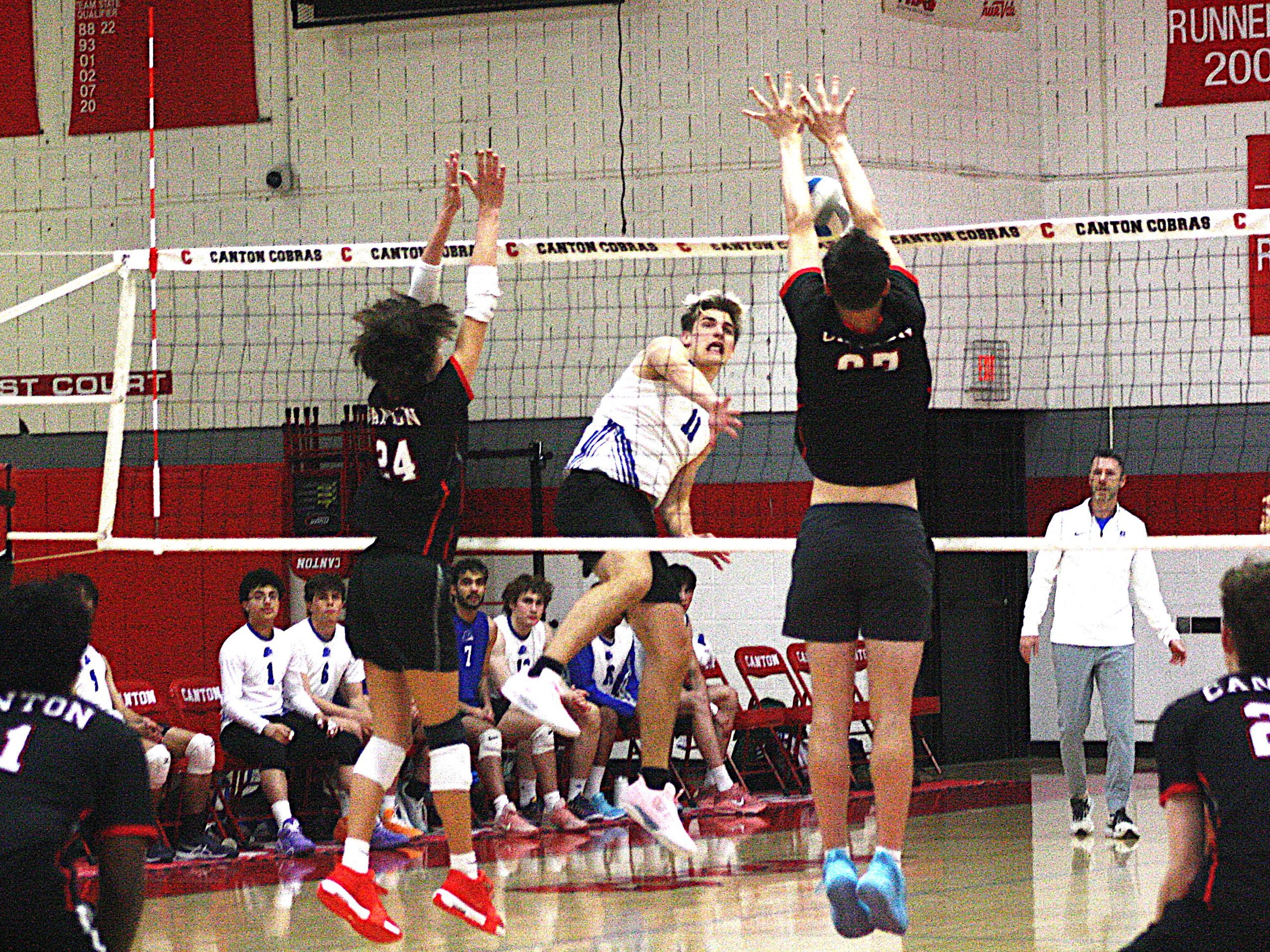 Volleyball players jump at the net during a match as teammates sit on the bench watching.
