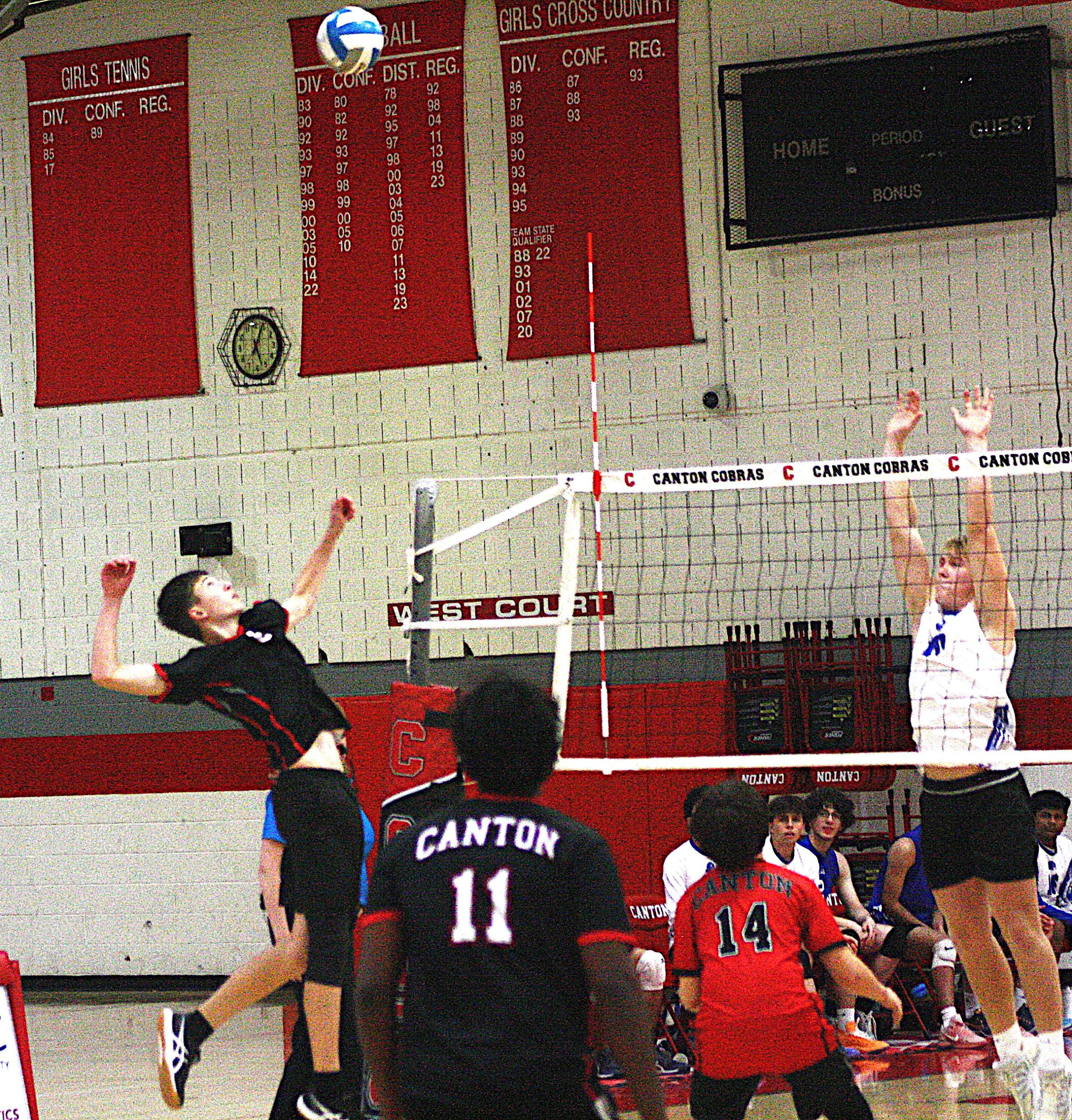 vbreg3 - Social House News Girls volleyball game in a gym as players in red uniforms jump near the net while teammates cheer nearby, Canton visible on jerseys and banners.