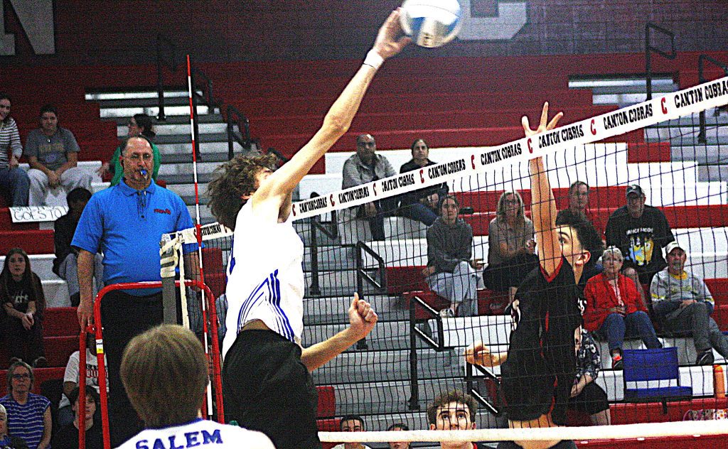 Volleyball player leaps to spike the ball over the net as spectators watch from red bleachers in a gymnasium.