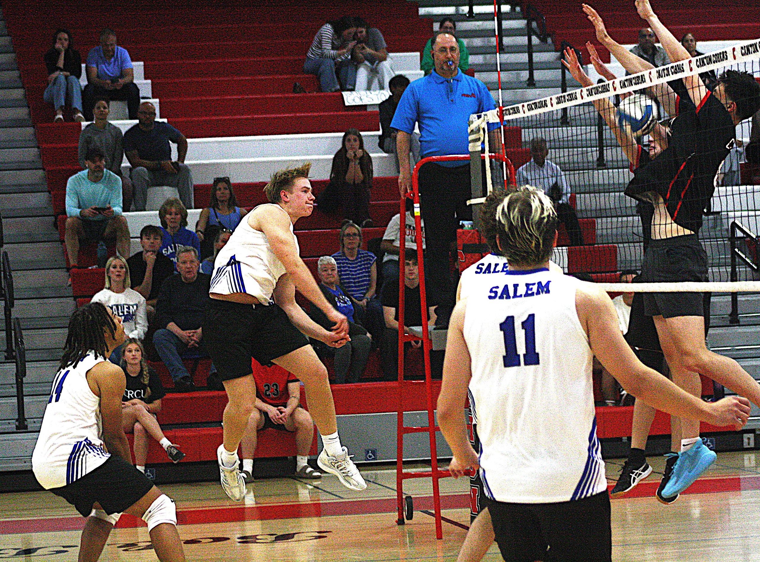 vbreg7 - Social House News Volleyball game in a gym: Salem players in white uniforms near the net while a referee blows a whistle on a red stand and spectators watch from red bleachers.