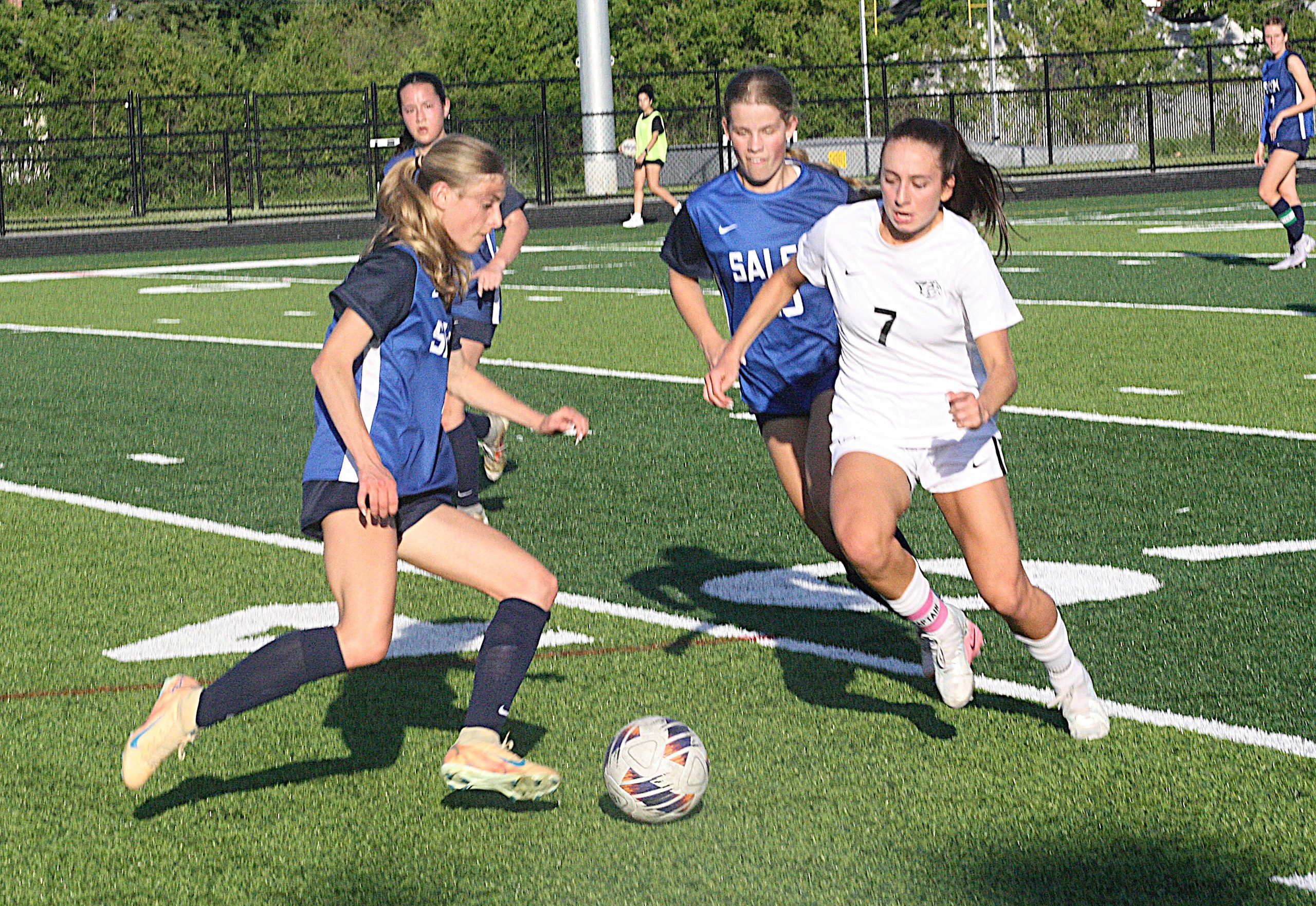 Girls' soccer match on a turf field; players in blue and white uniforms chase the ball, close-up action shot on the field surface with a goal line nearby.