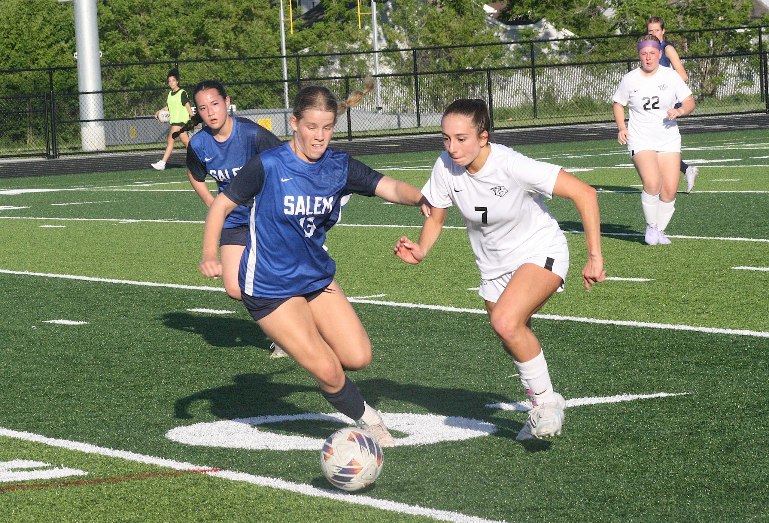 Girls' soccer match: two players in blue Salem jerseys and white jerseys chase the ball on a turf field, foreground action near the goal line.