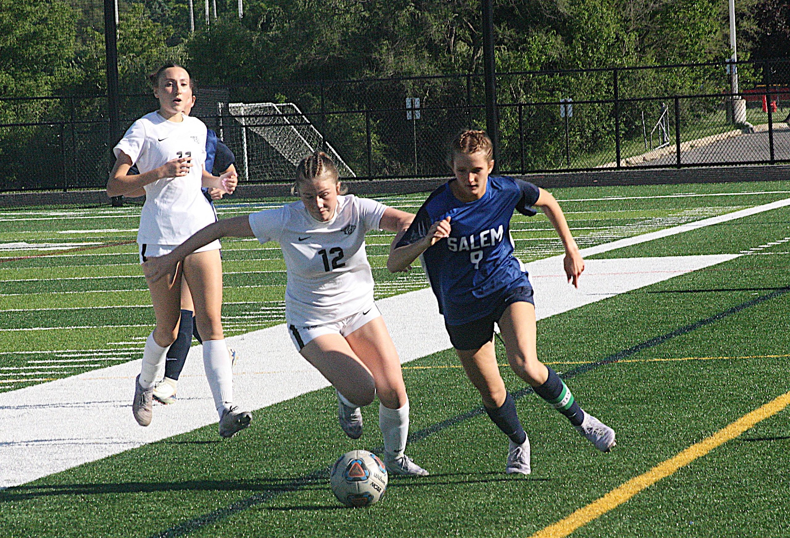Three female soccer players chase a ball on an artificial turf field, two in white uniforms and one in a blue Salem jersey.
