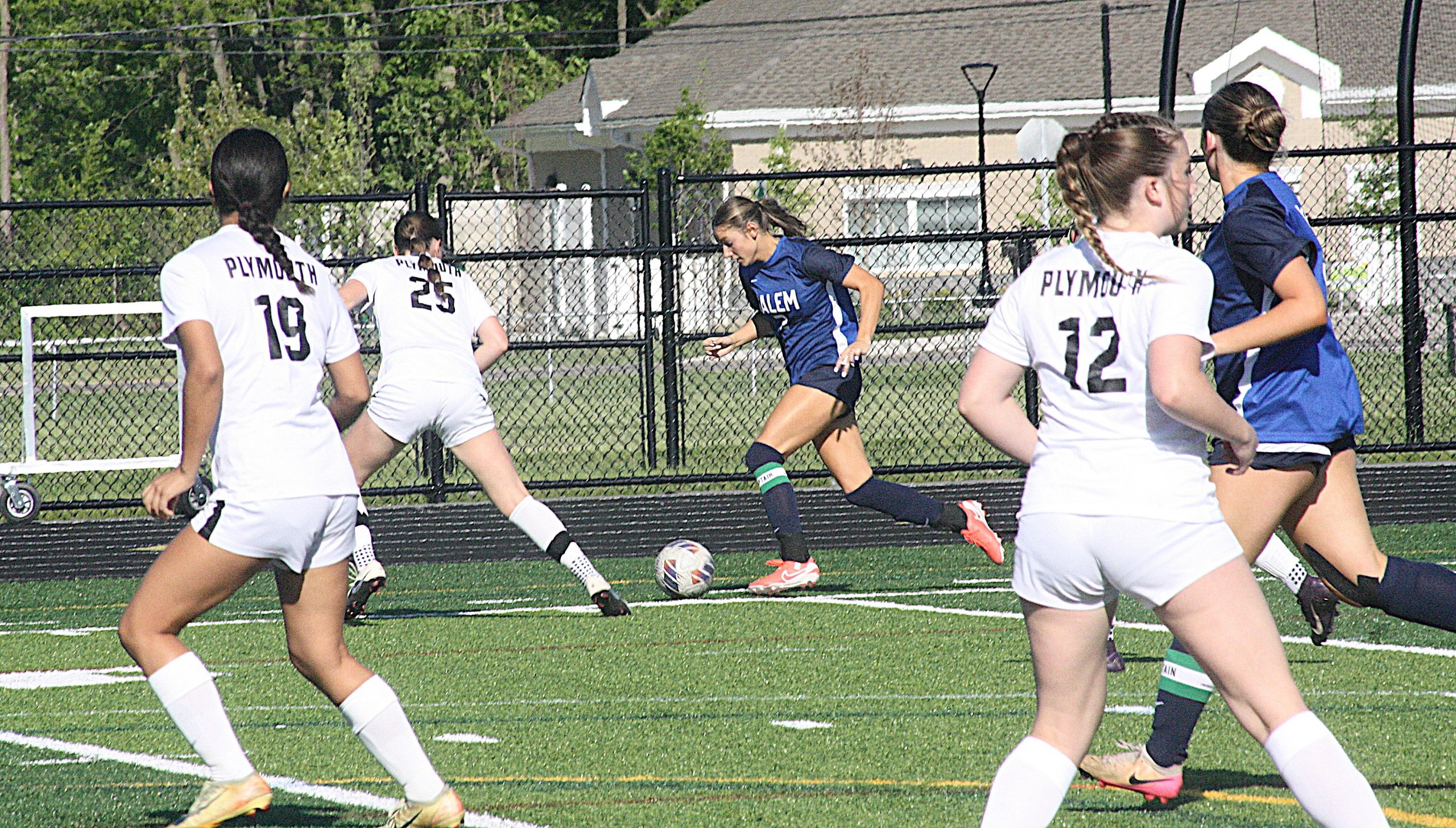Girls' soccer game in progress on artificial turf; white Plymouth uniforms chase a ball while a blue-clad opponent runs nearby near a chain-link fence.