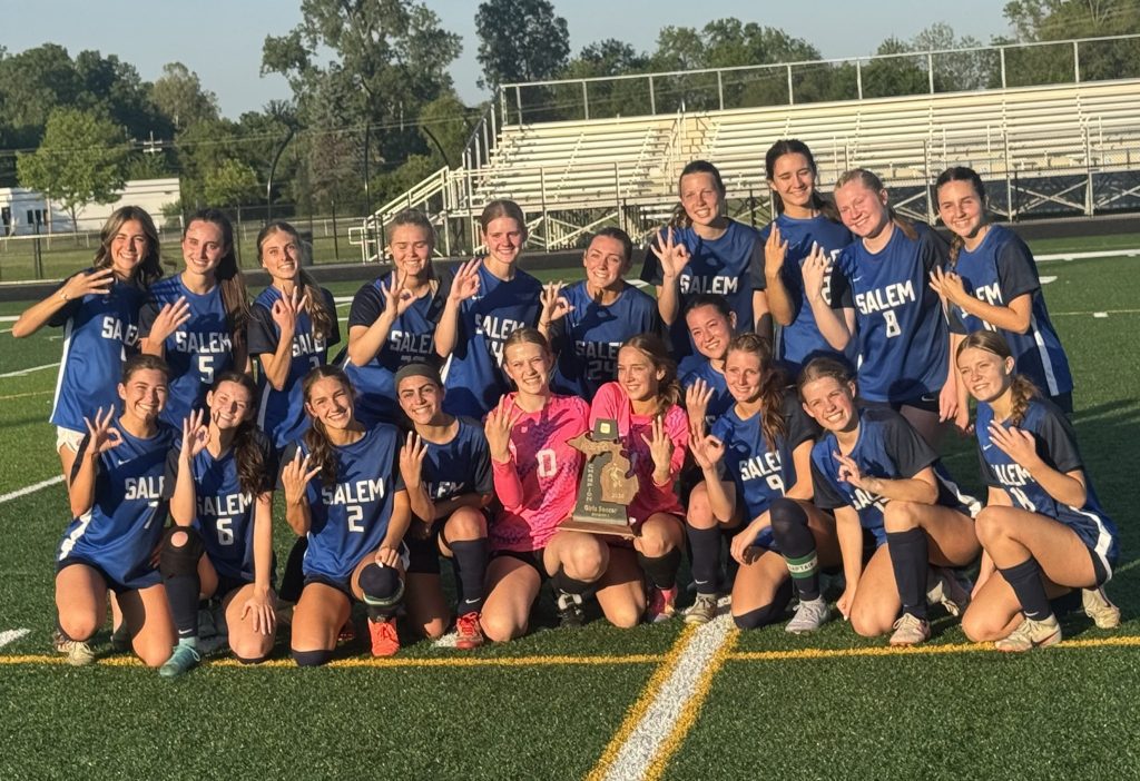 Salem girls soccer team posing on the field with trophy after a win, wearing blue jerseys and making hand signs.