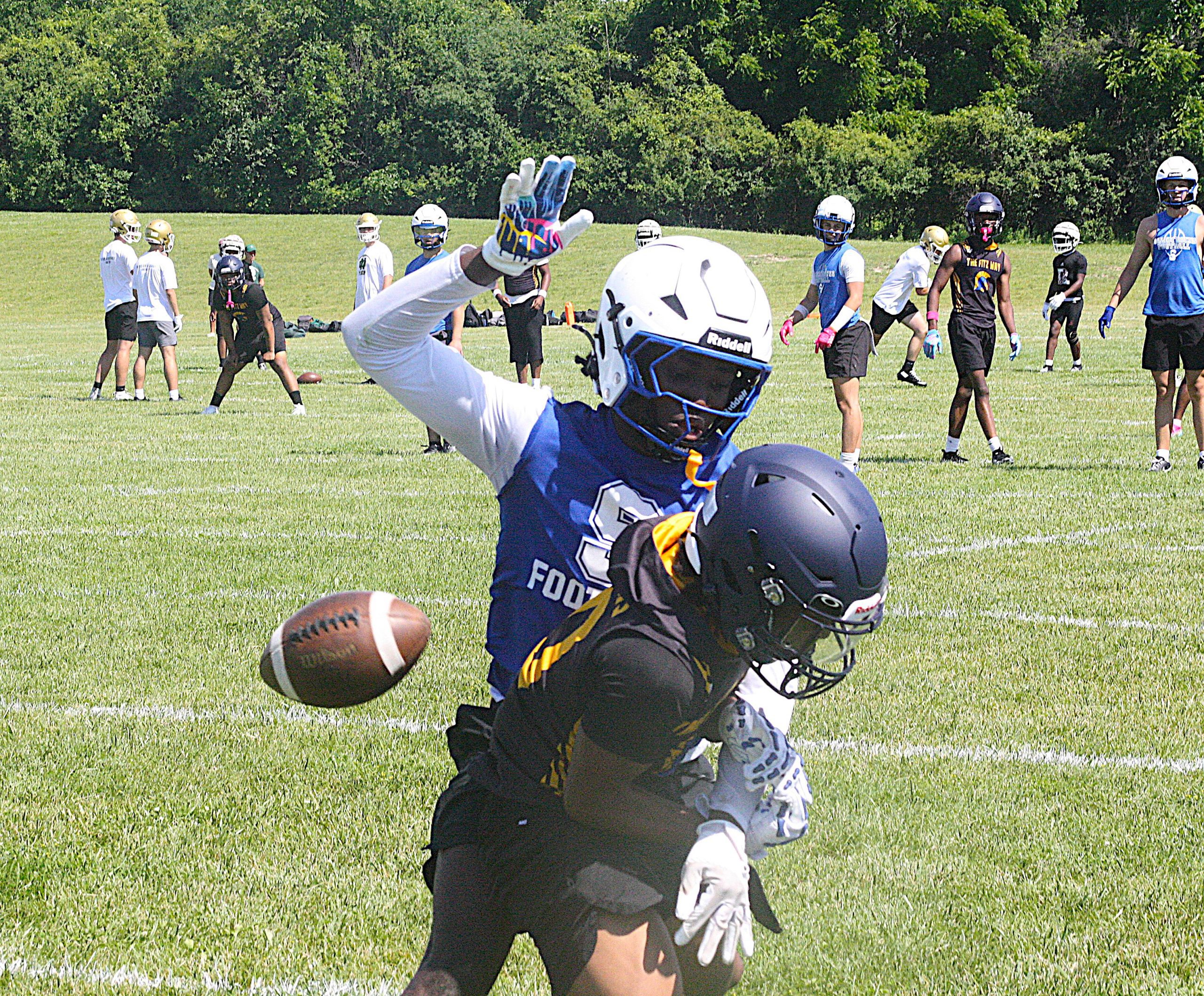 Football players wearing helmets and pads in a practice on a grassy field, with a ball rolling toward the foreground and a blue-and-white jerseyed player in the foreground reaching for it.