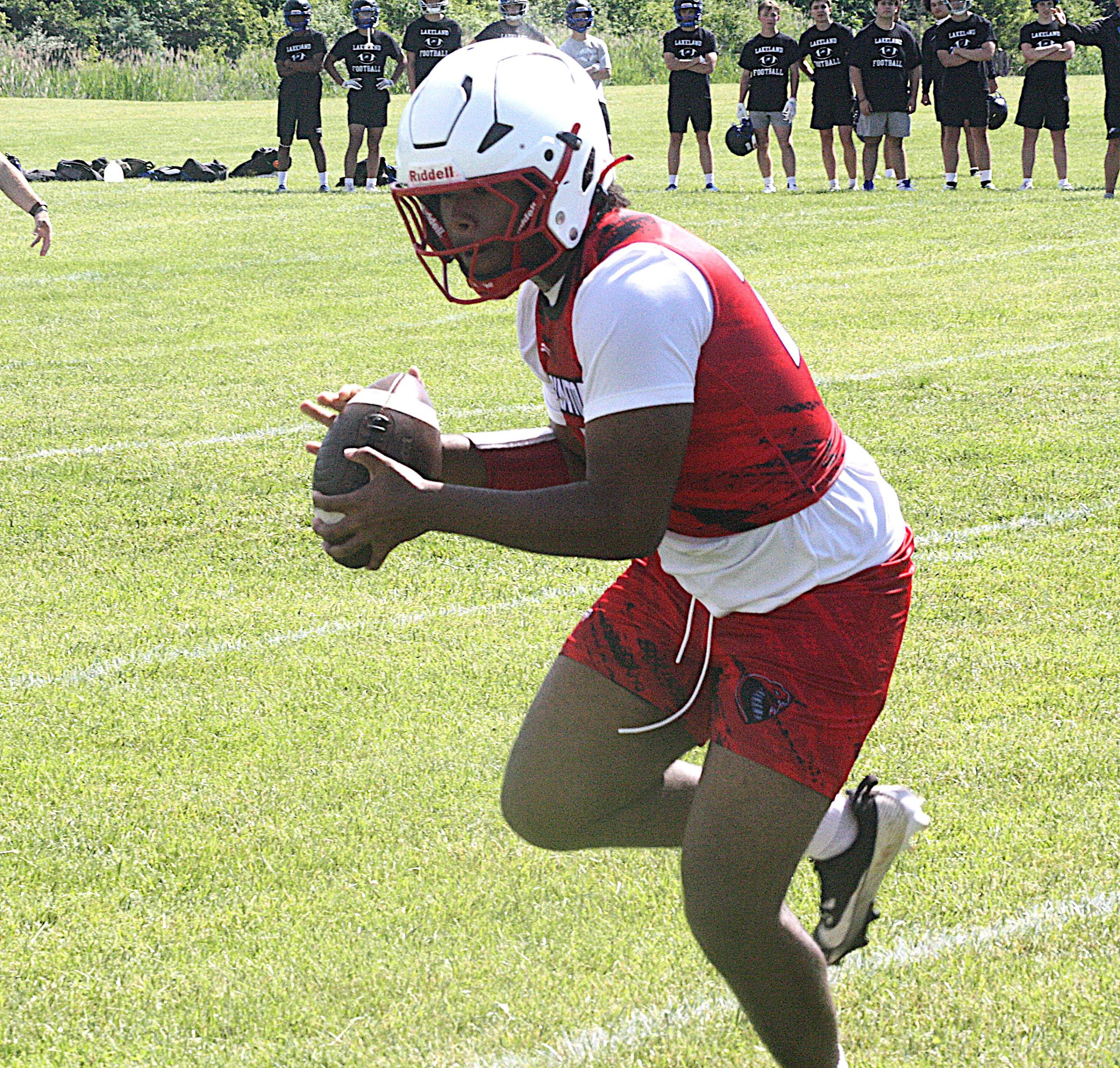 Football player in red and white uniform lunges with the ball toward the ground on a grassy field, with a line of teammates in dark shirts watching in the background.