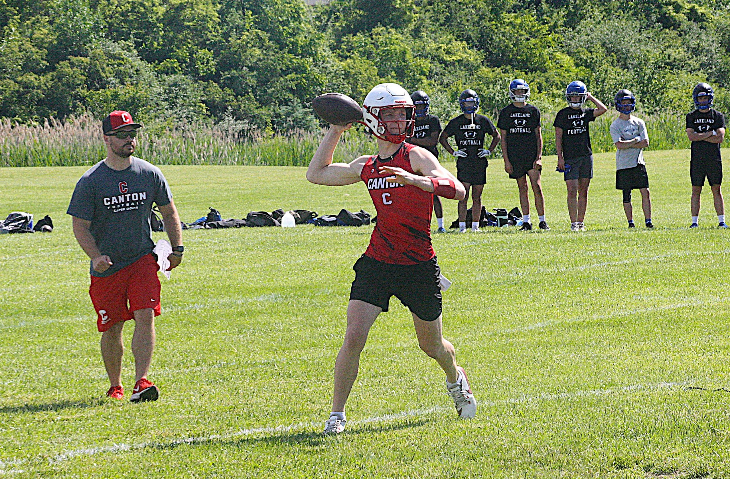 Young football player in a red Canton jersey throws a pass as teammates in black Lakeland shirts watch on a sunny grassy field.