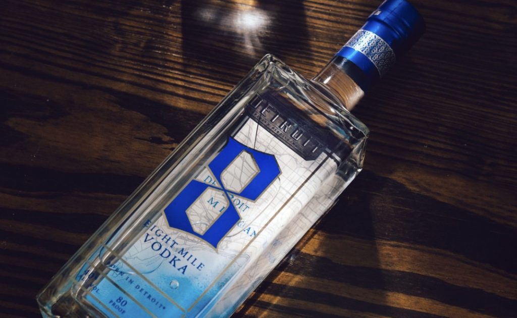 Clear rectangular vodka bottle with a blue and silver label on a dark wooden surface, cap sealed with blue foil (80 proof visible on the label).