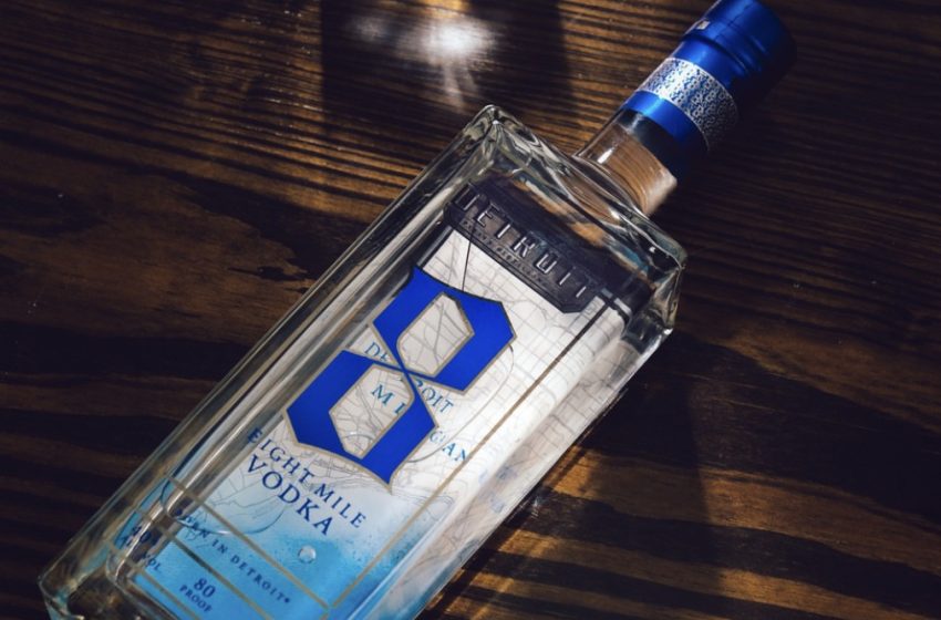 Clear rectangular vodka bottle with a blue and silver label on a dark wooden surface, cap sealed with blue foil (80 proof visible on the label).