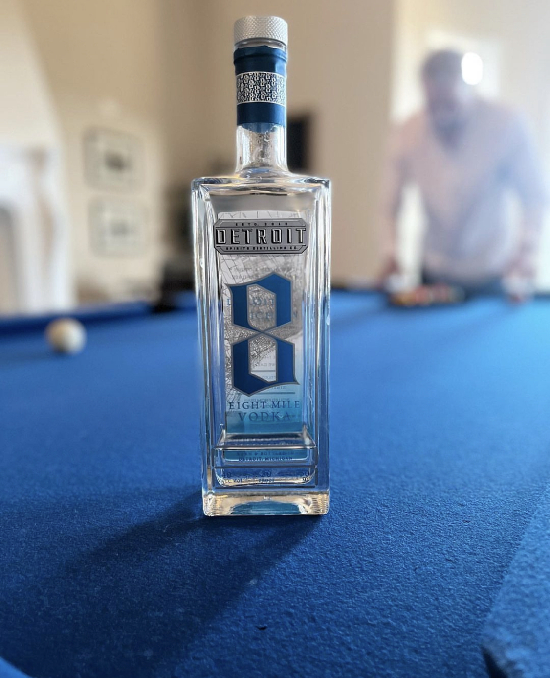 Bottle of Detroit Eight Mile Vodka on a blue pool table with a blurred figure in the background.