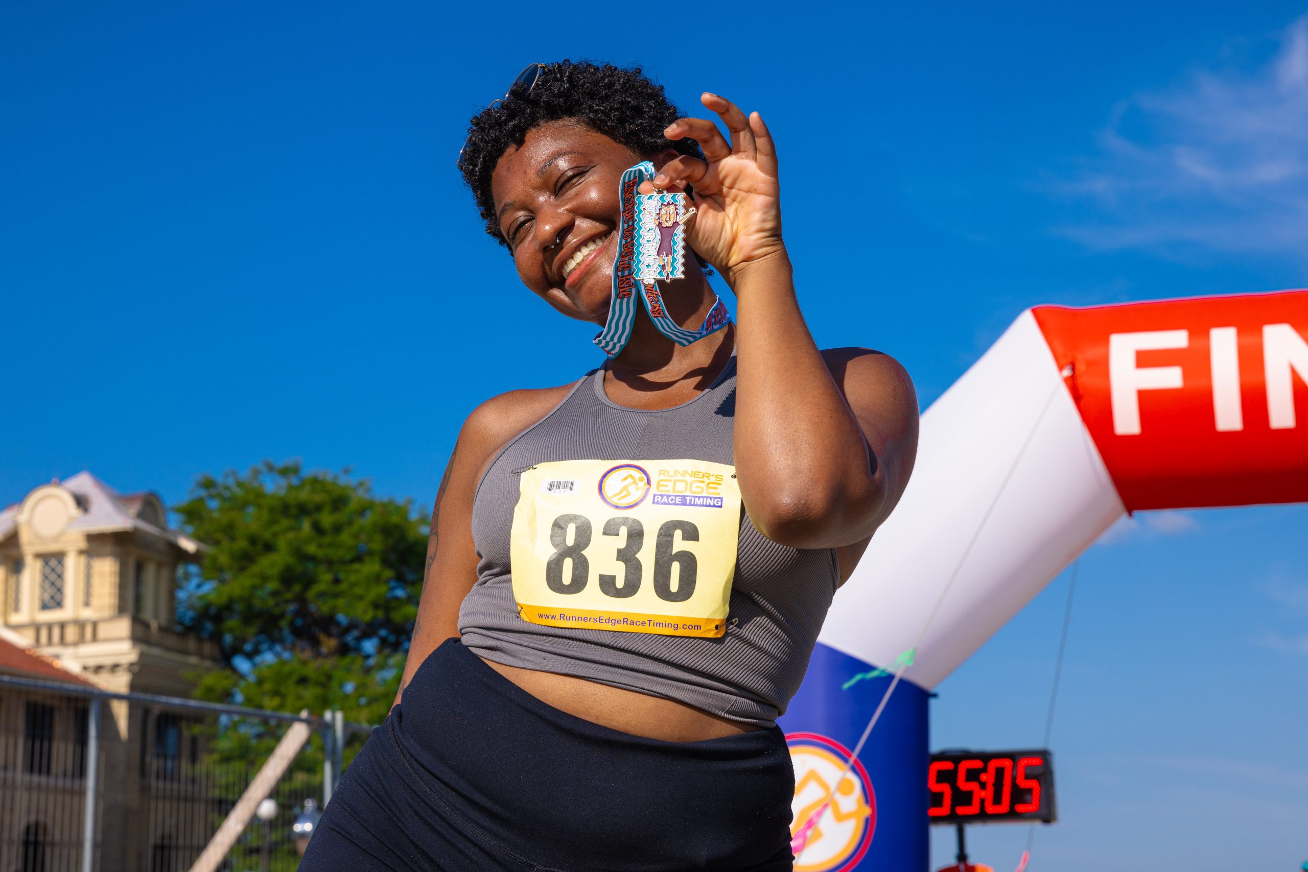 Smiling runner holds up a finish-line medal beside a blue sky, bib number 836 visible on gray top at a race finish.