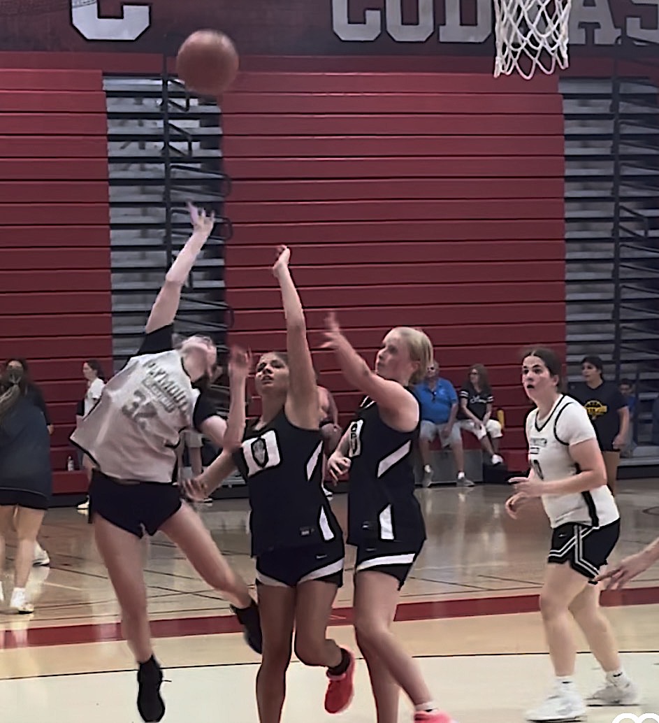 Screenshot - Social House News Girls' basketball game: three players reach for the ball near the basket as it hangs in midair, with red bleachers in the background.